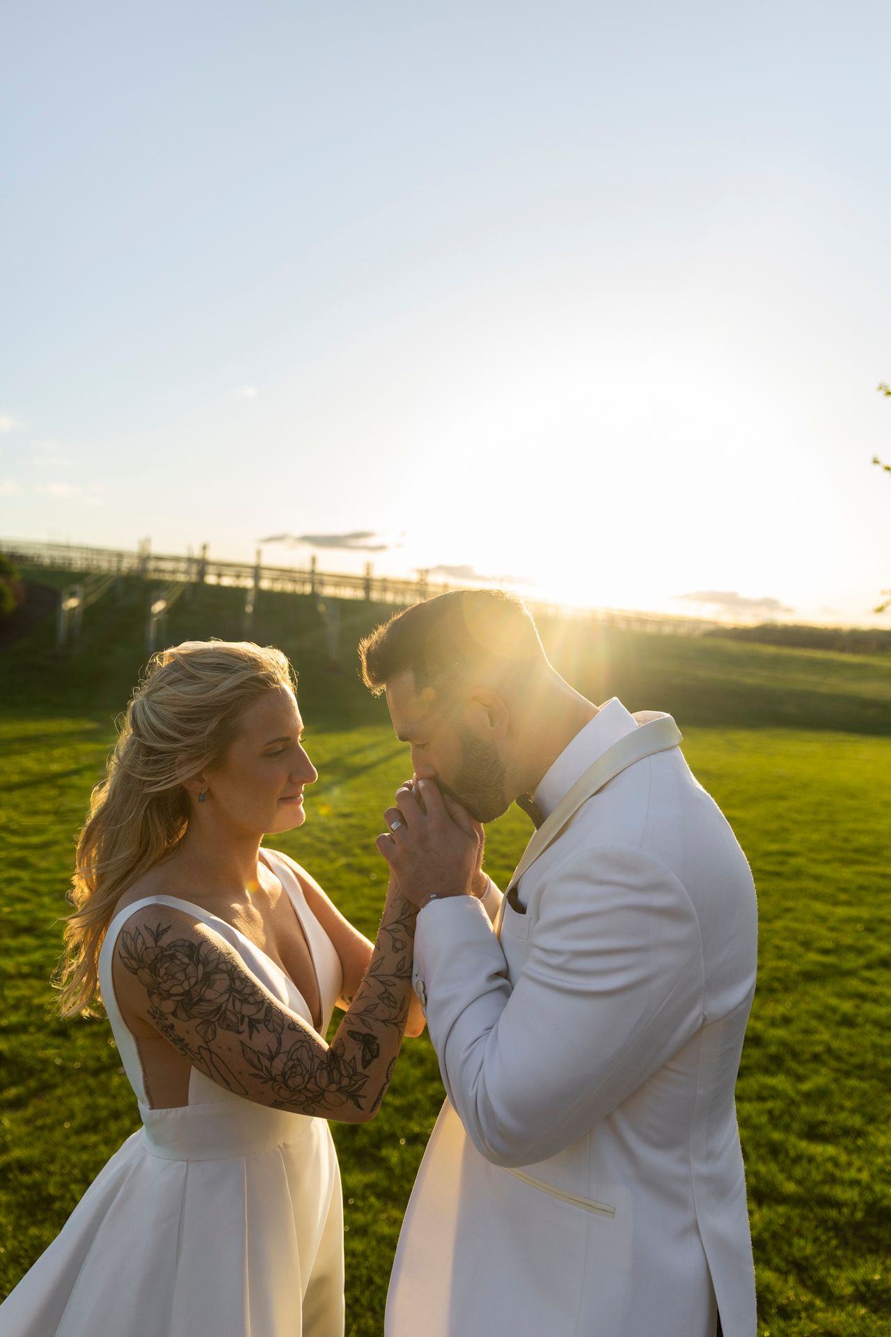 Groom kisses bride's hand at golden sunset on lush green lawn at outdoor wedding.