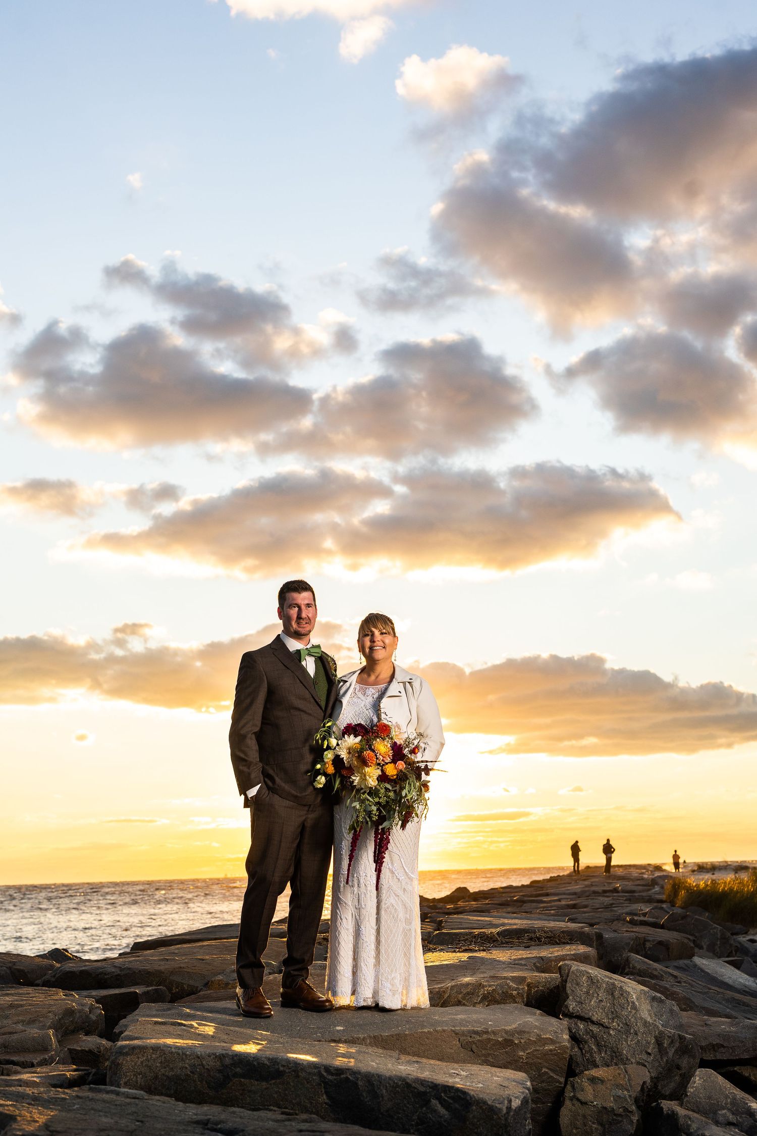 Bride and groom posing on rocky jetty at vibrant golden sunset with colorful floral bouquet.