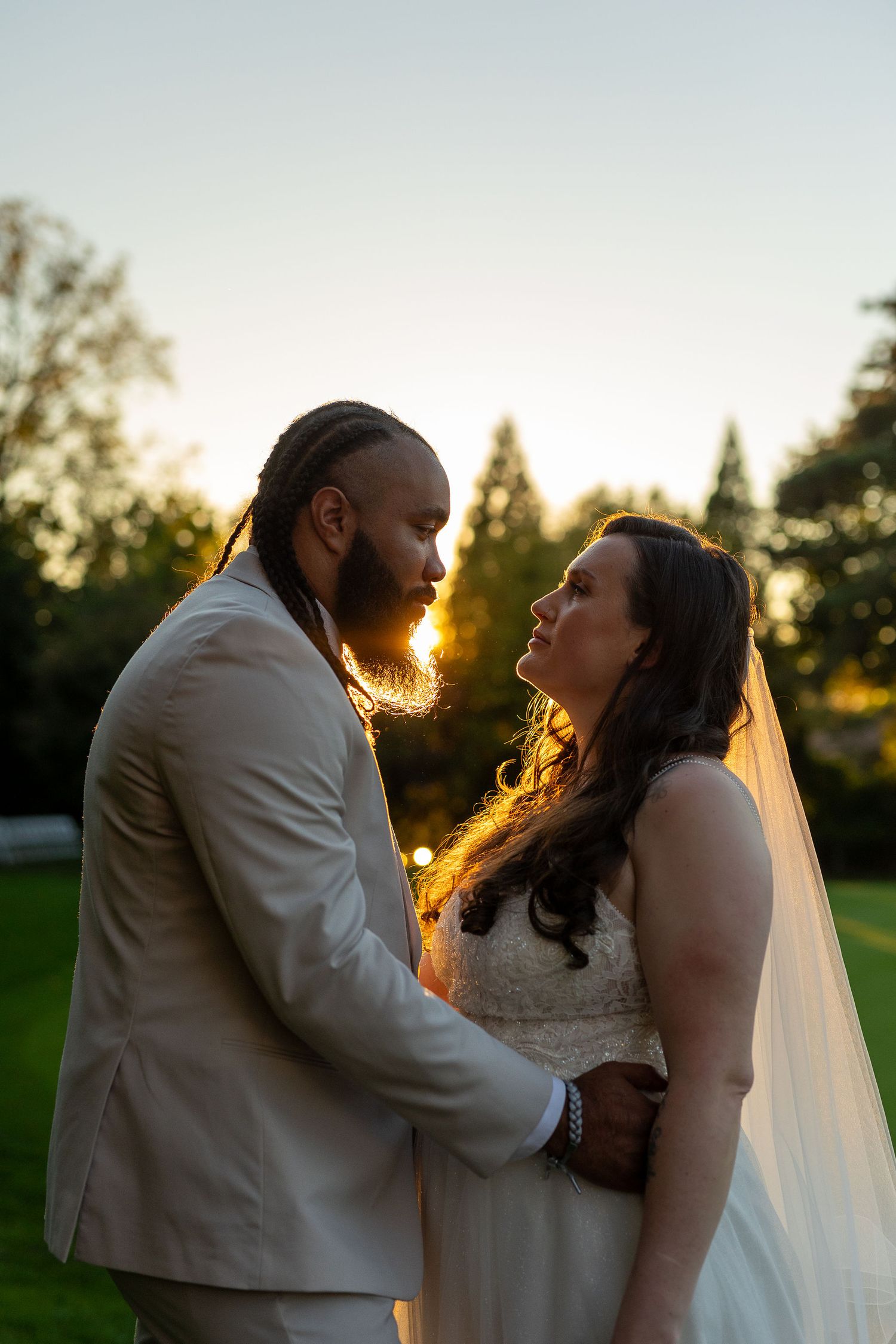 Bride gazes up at groom during golden hour sunset, both embracing in a romantic outdoor wedding portrait.