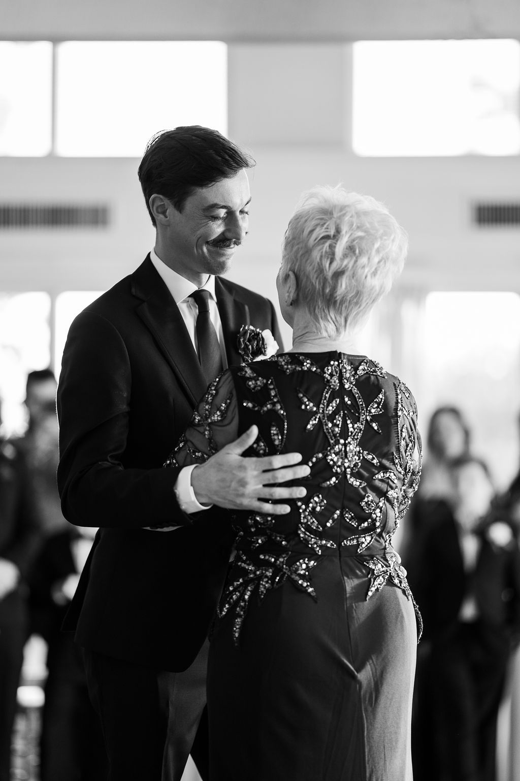 Groom in black suit shares tender mother-son dance at elegant wedding reception, black and white.