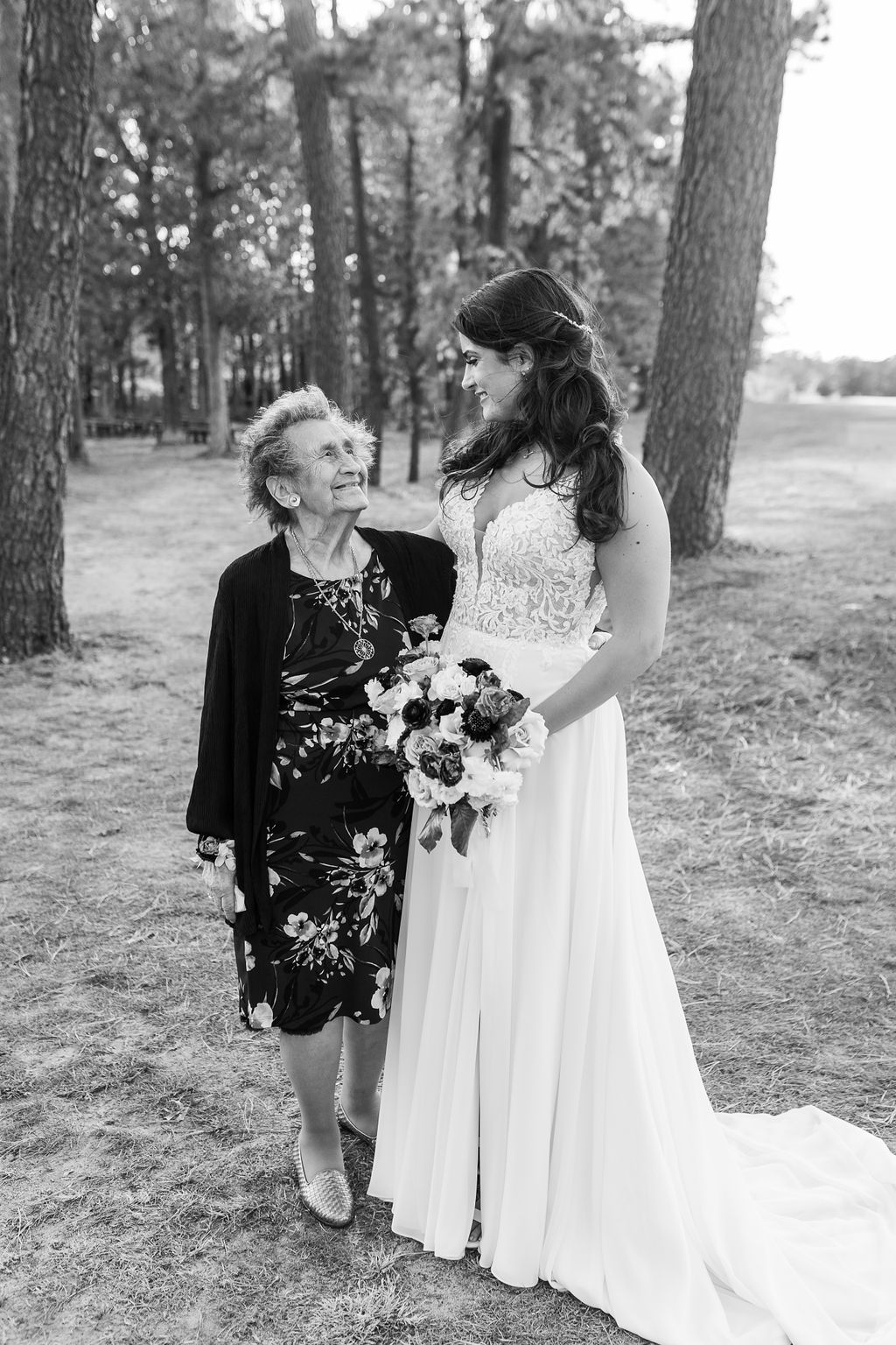 Bride in white lace gown holds bouquet while posing with elderly woman in black floral dress outdoors.