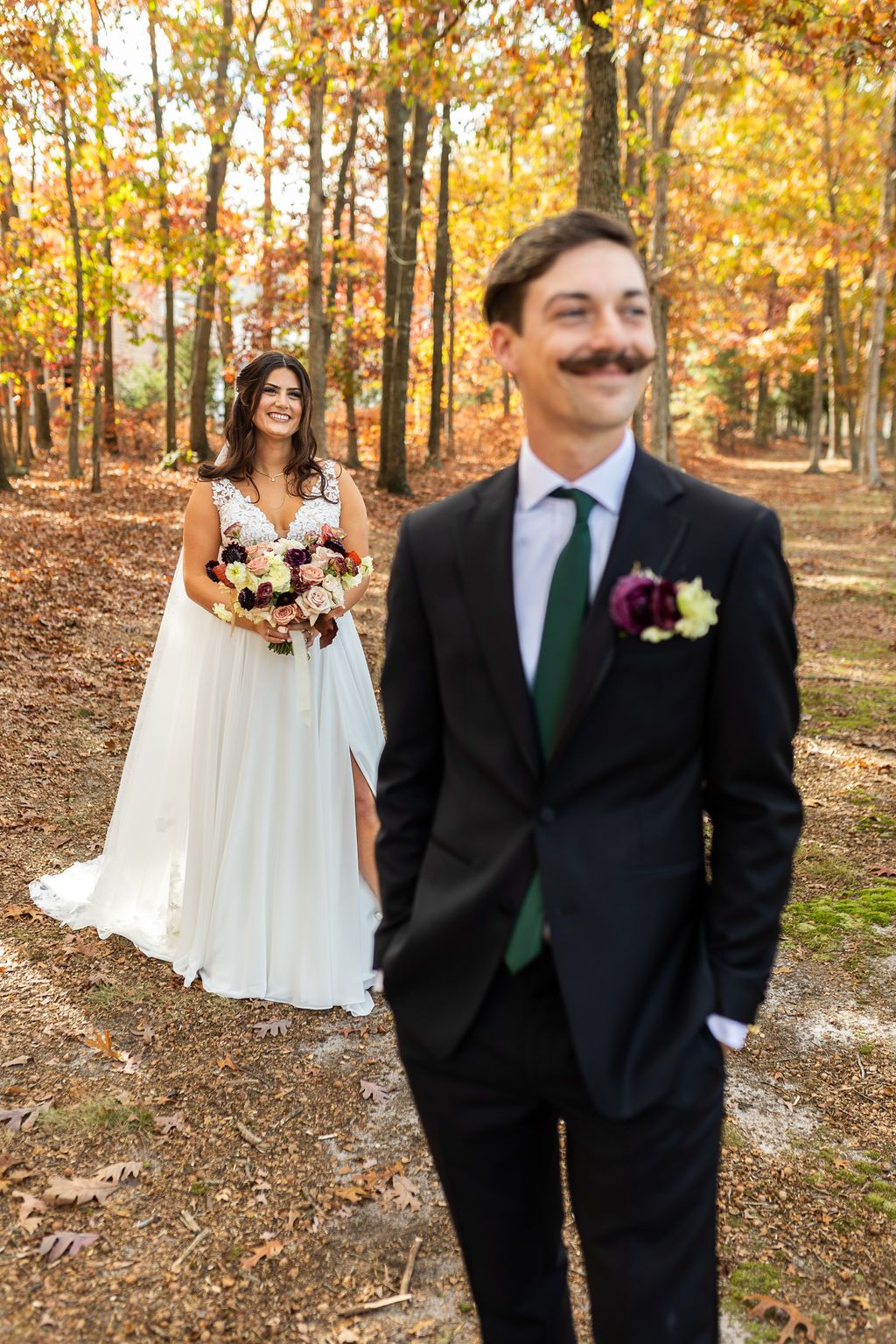 Groom with mustache stands in autumn forest while bride smiles behind him.