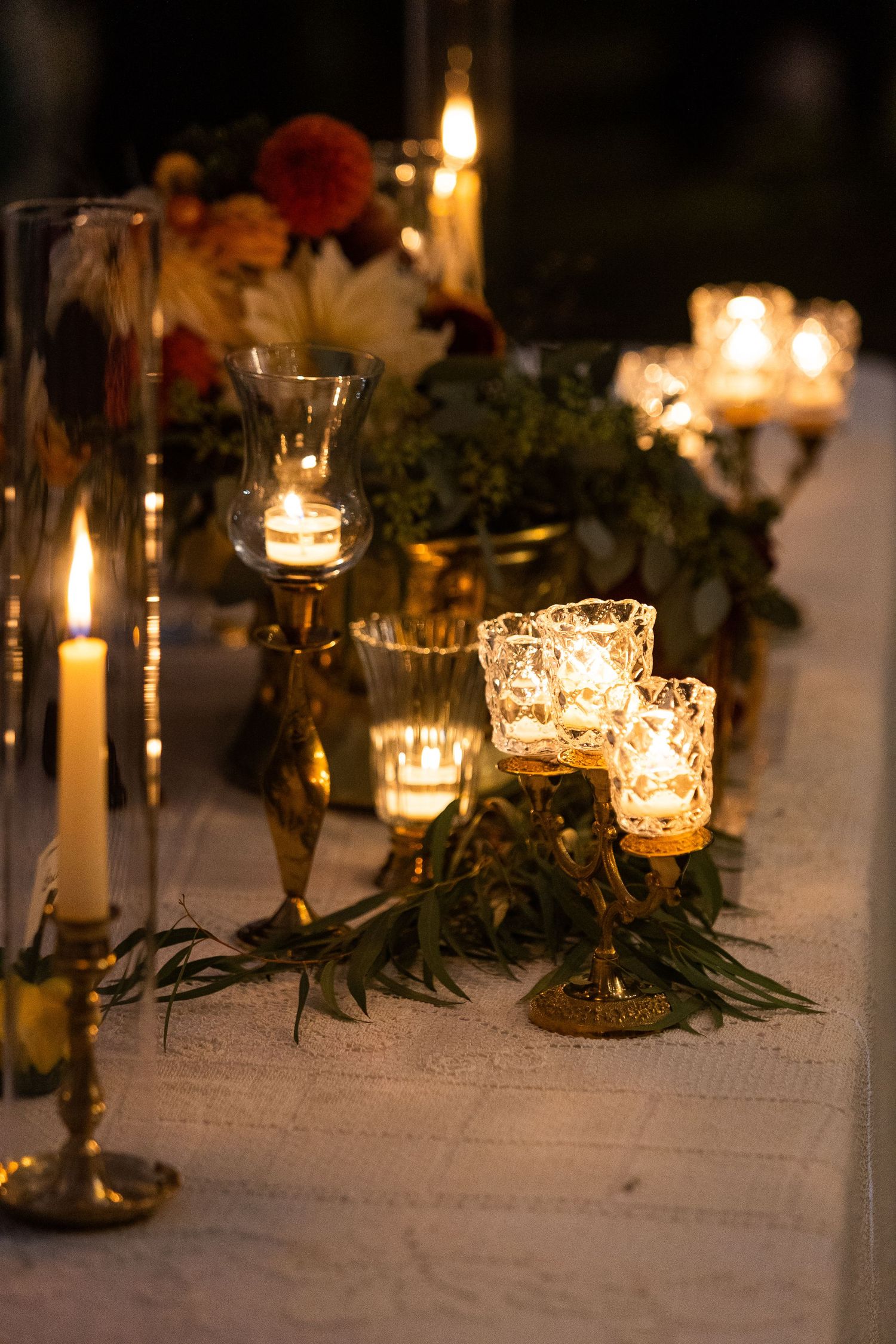 Elegant wedding table decorated with glowing candles, gold candleholders, and lush greenery at night.