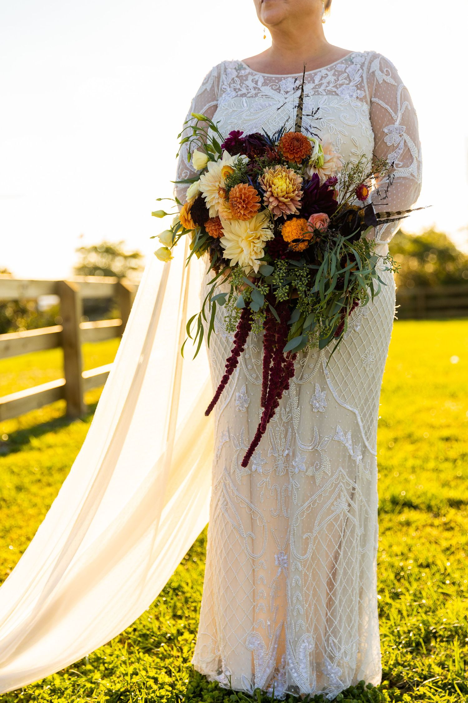 Bride in beaded lace gown holding lush cascading bouquet of dahlias and amaranthus in warm sunset light.