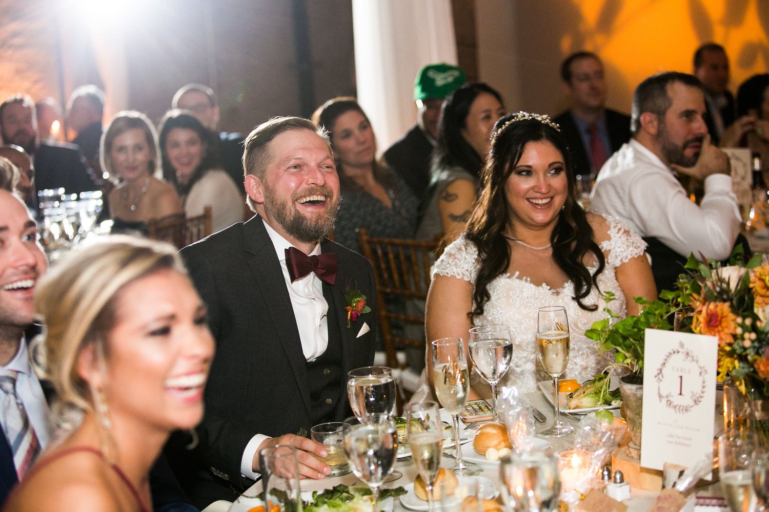 A laughing bride and groom in formal attire sit at their wedding reception dinner table.