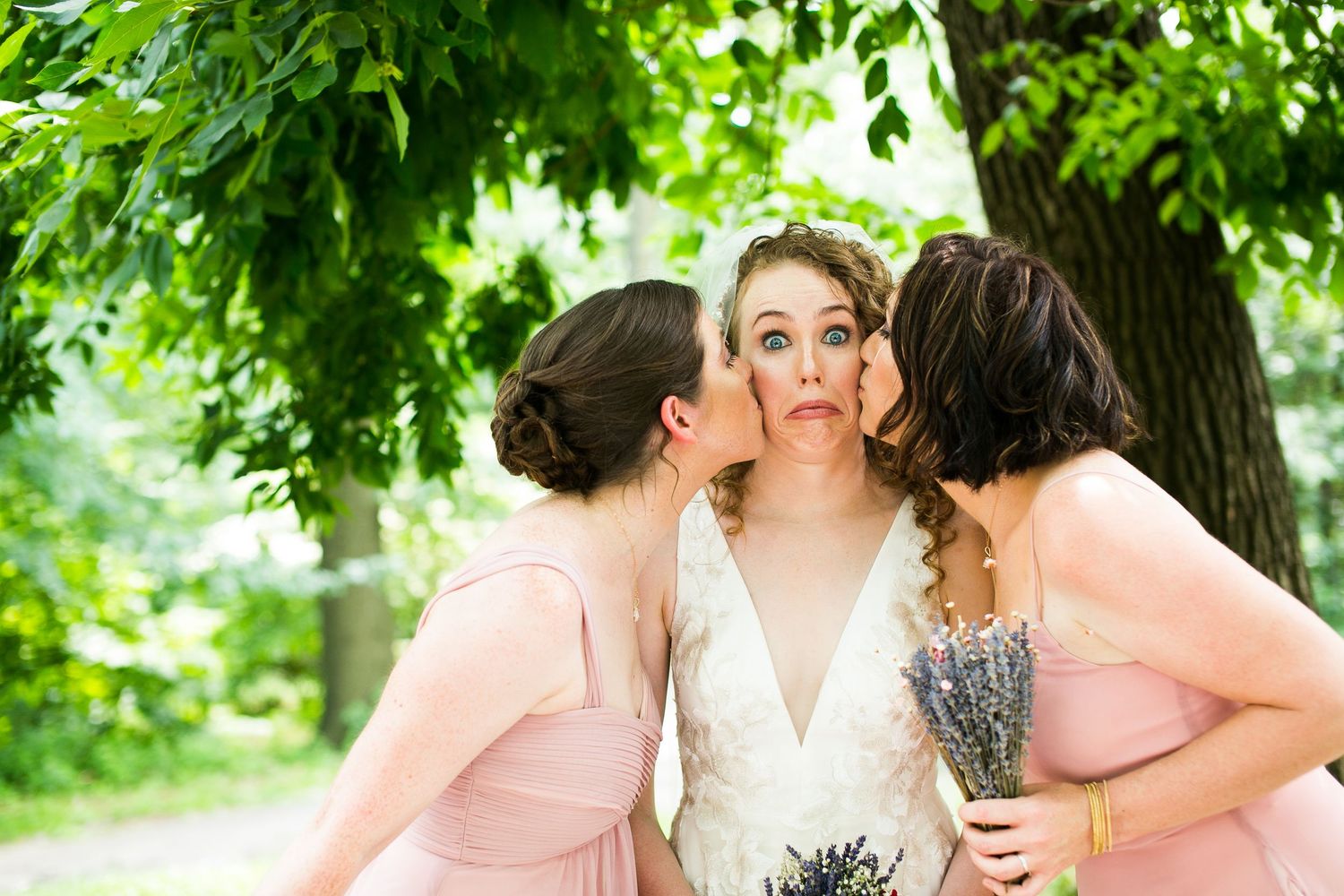 Bride makes funny face while two bridesmaids in pink dresses kiss her cheeks outdoors.