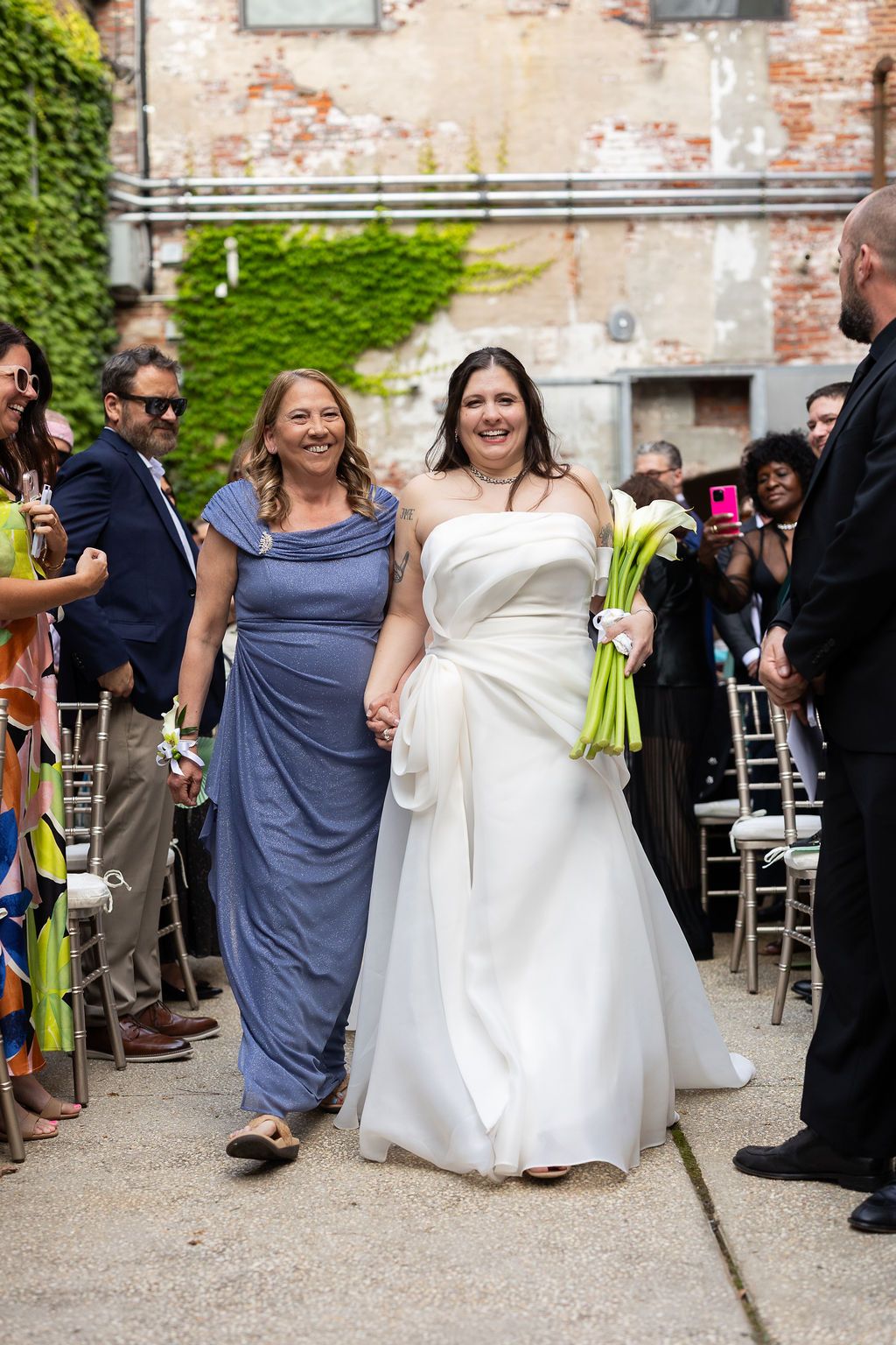 Smiling bride in strapless white gown walks down aisle with woman in blue dress at outdoor wedding.