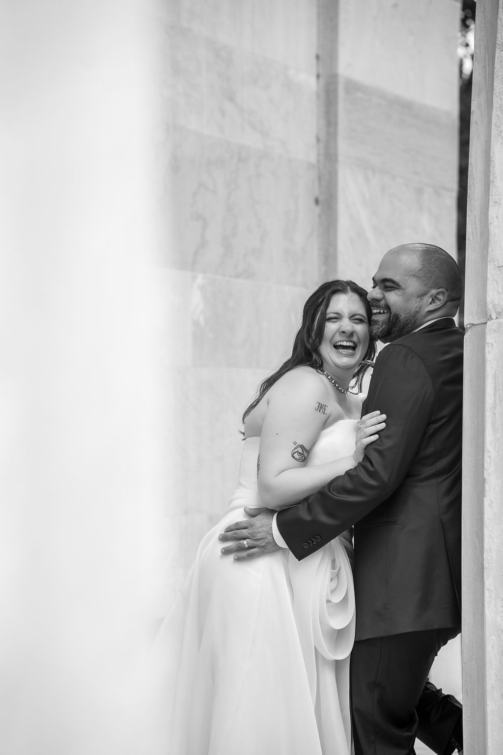 Groom embracing laughing bride against marble wall in a joyful black and white wedding photo.
