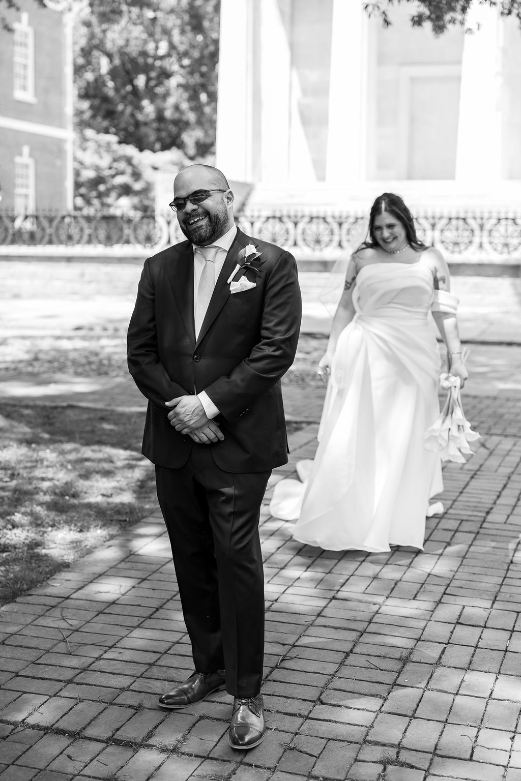 Black and white first look photo of groom in dark suit awaiting bride approaching on brick path.