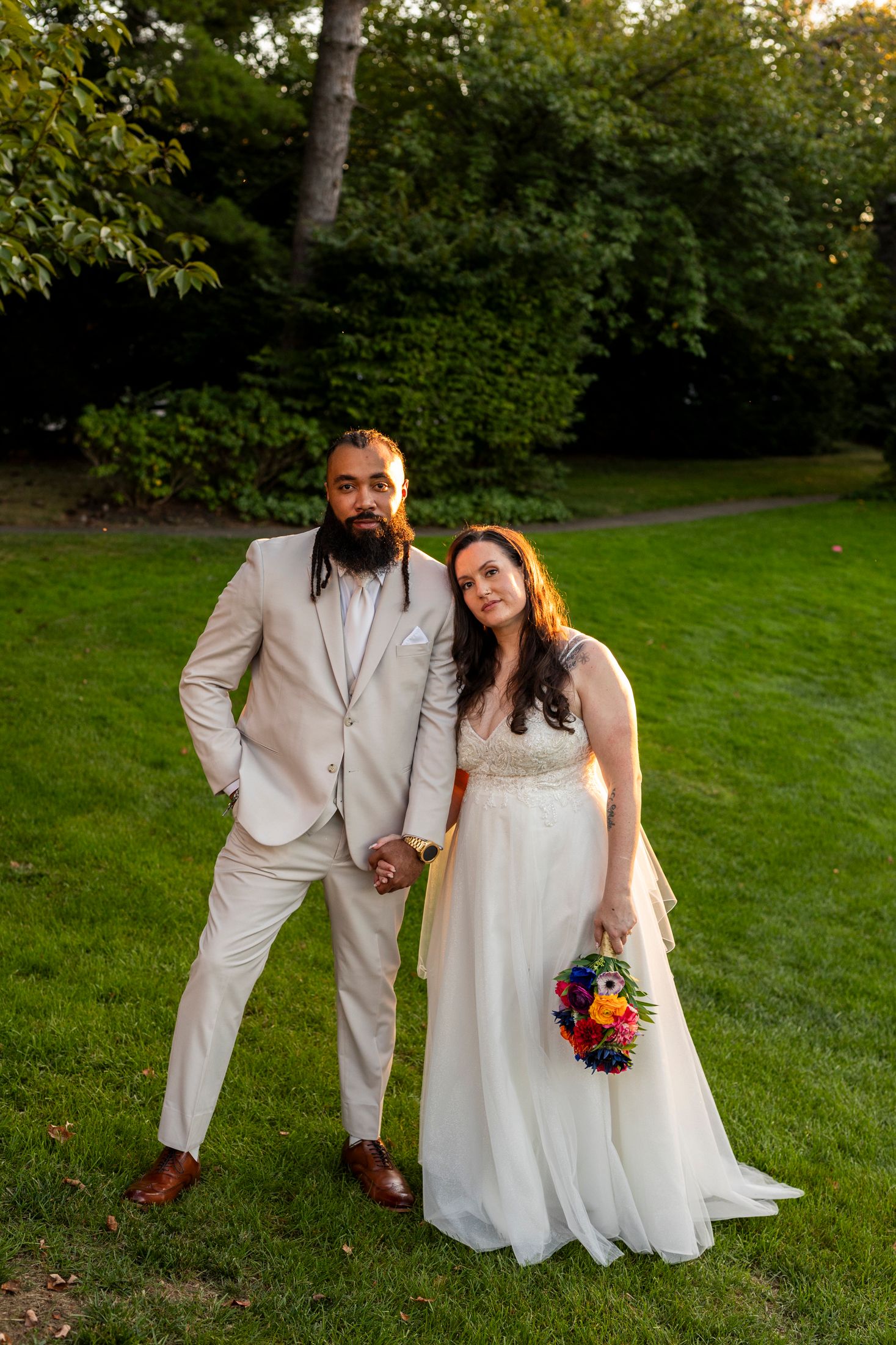 Bride and groom pose on green lawn at golden hour, bride holding colorful wildflower bouquet.