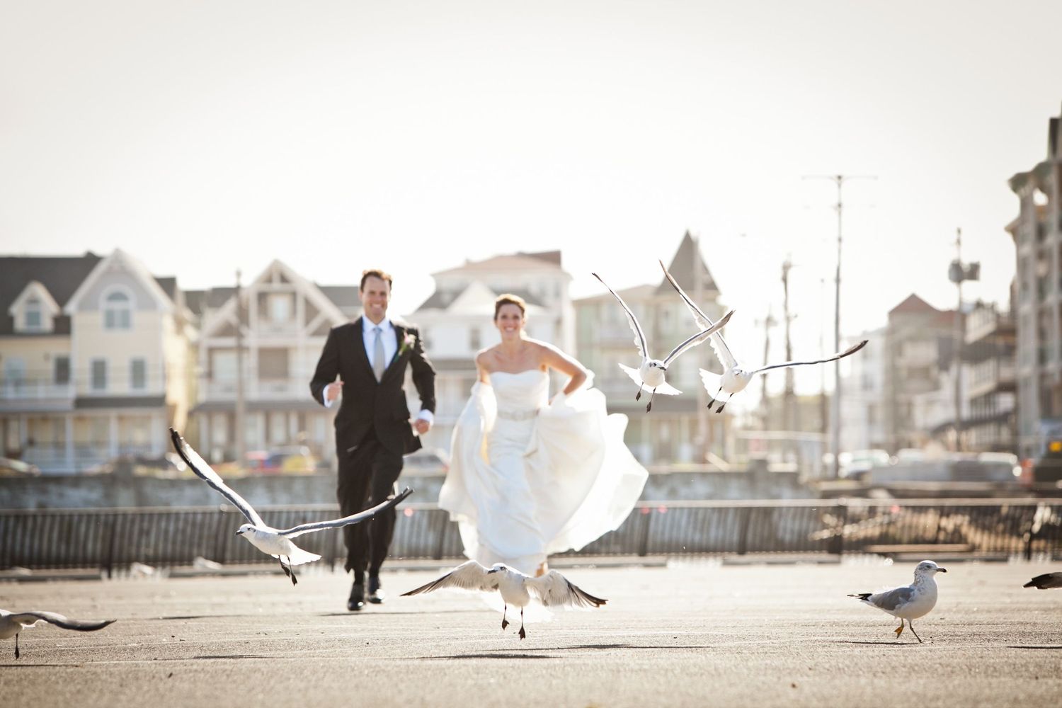 Bride and groom running on boardwalk with seagulls flying around them.