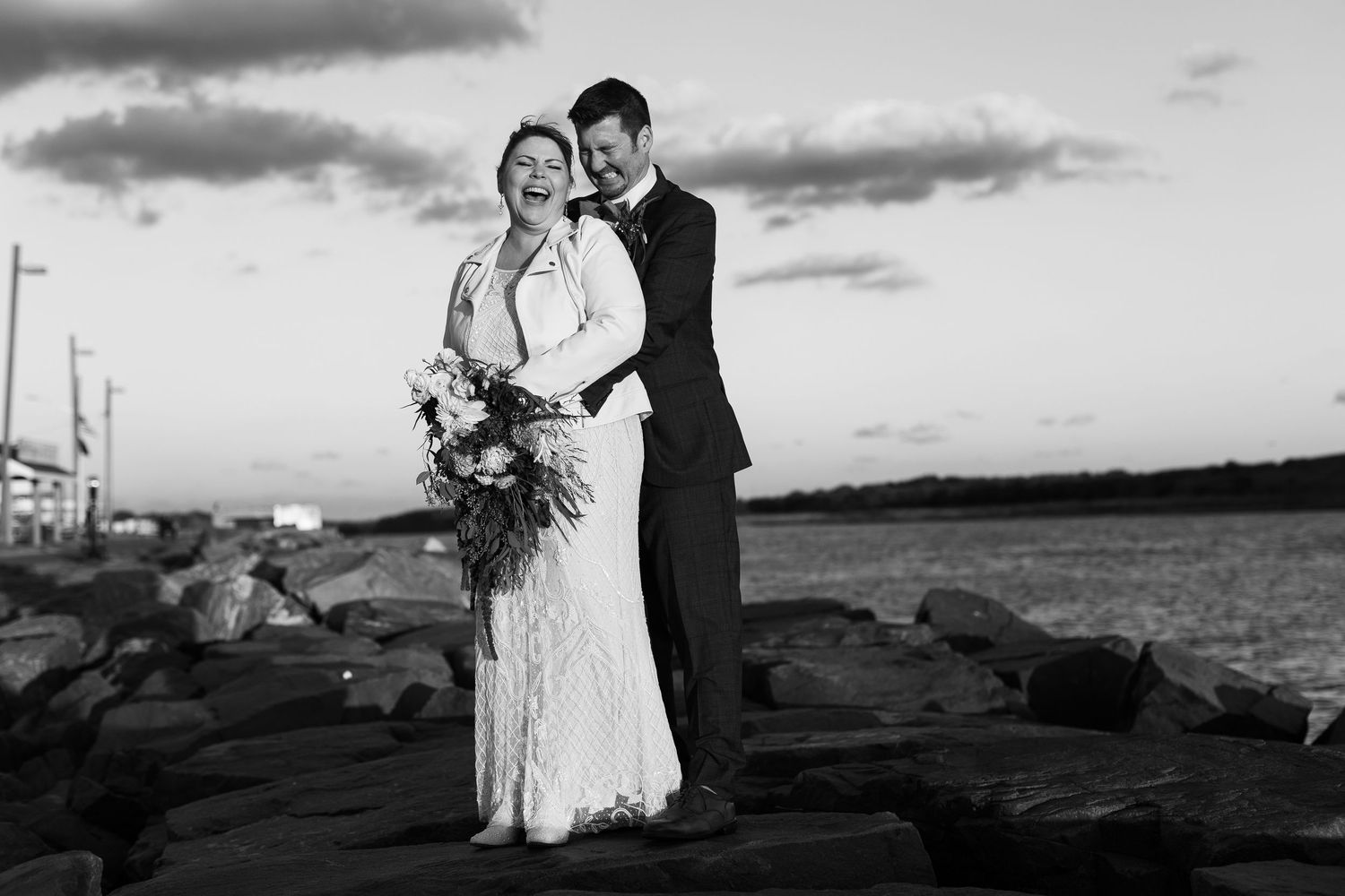 Laughing bride and groom share a joyful embrace on rocks by the sea at dusk.