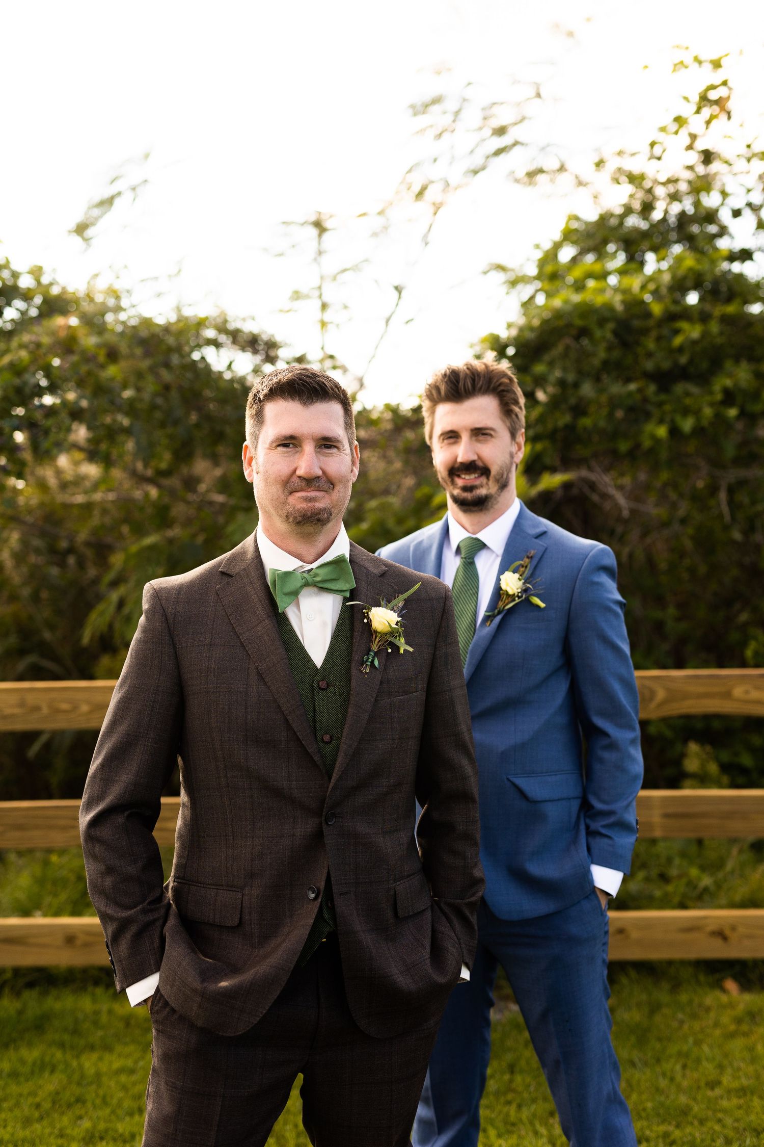 Two grooms in matching plaid suits with green bow ties and boutonnieres posing outdoors at wedding.