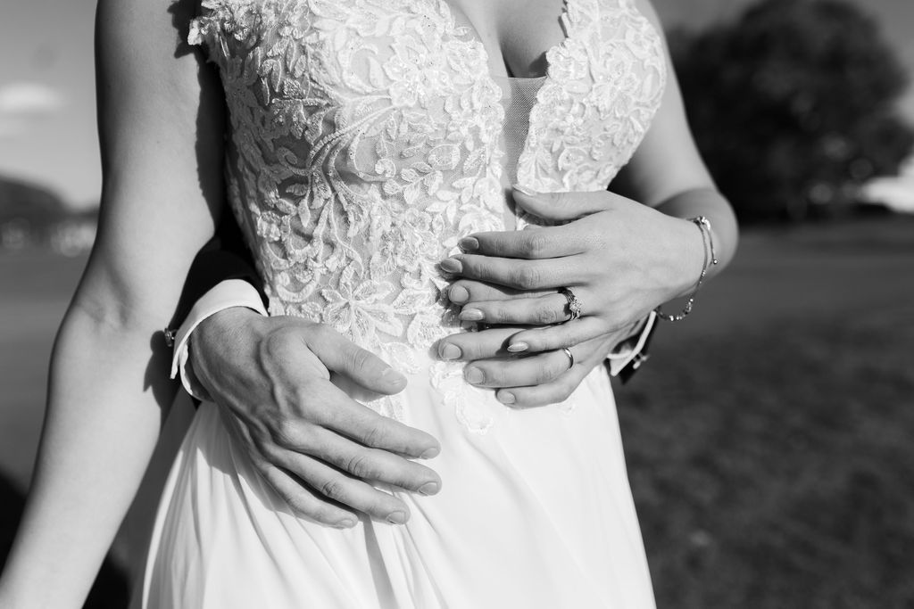Black and white close-up of groom embracing bride's waist in lace wedding gown, highlighting wedding rings.