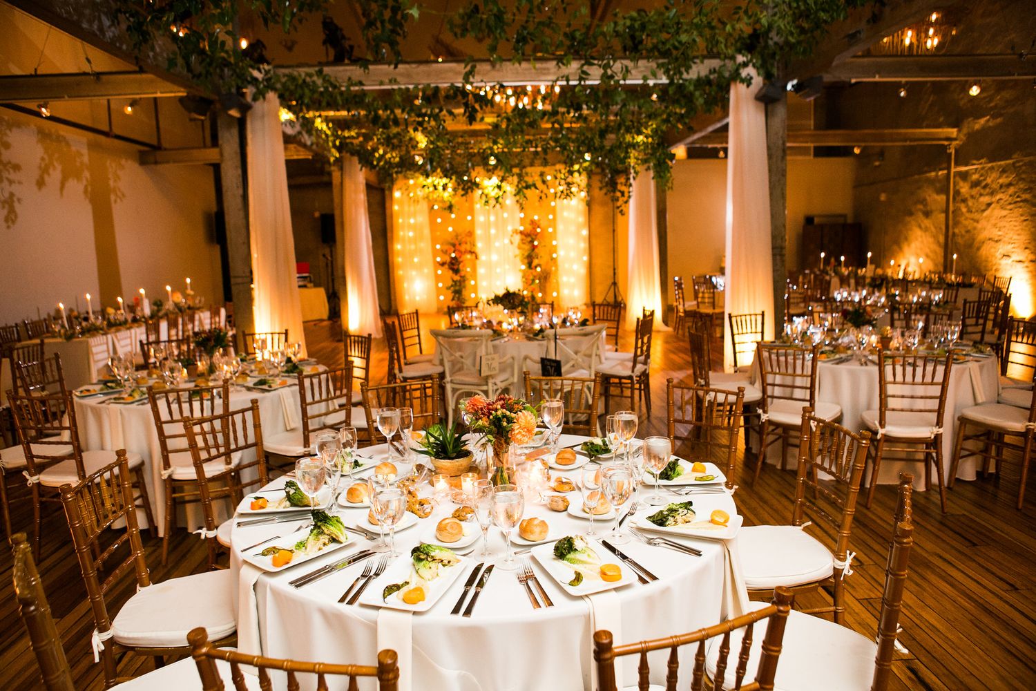 Warmly lit wedding reception hall with round tables, chiavari chairs, and hanging greenery overhead.
