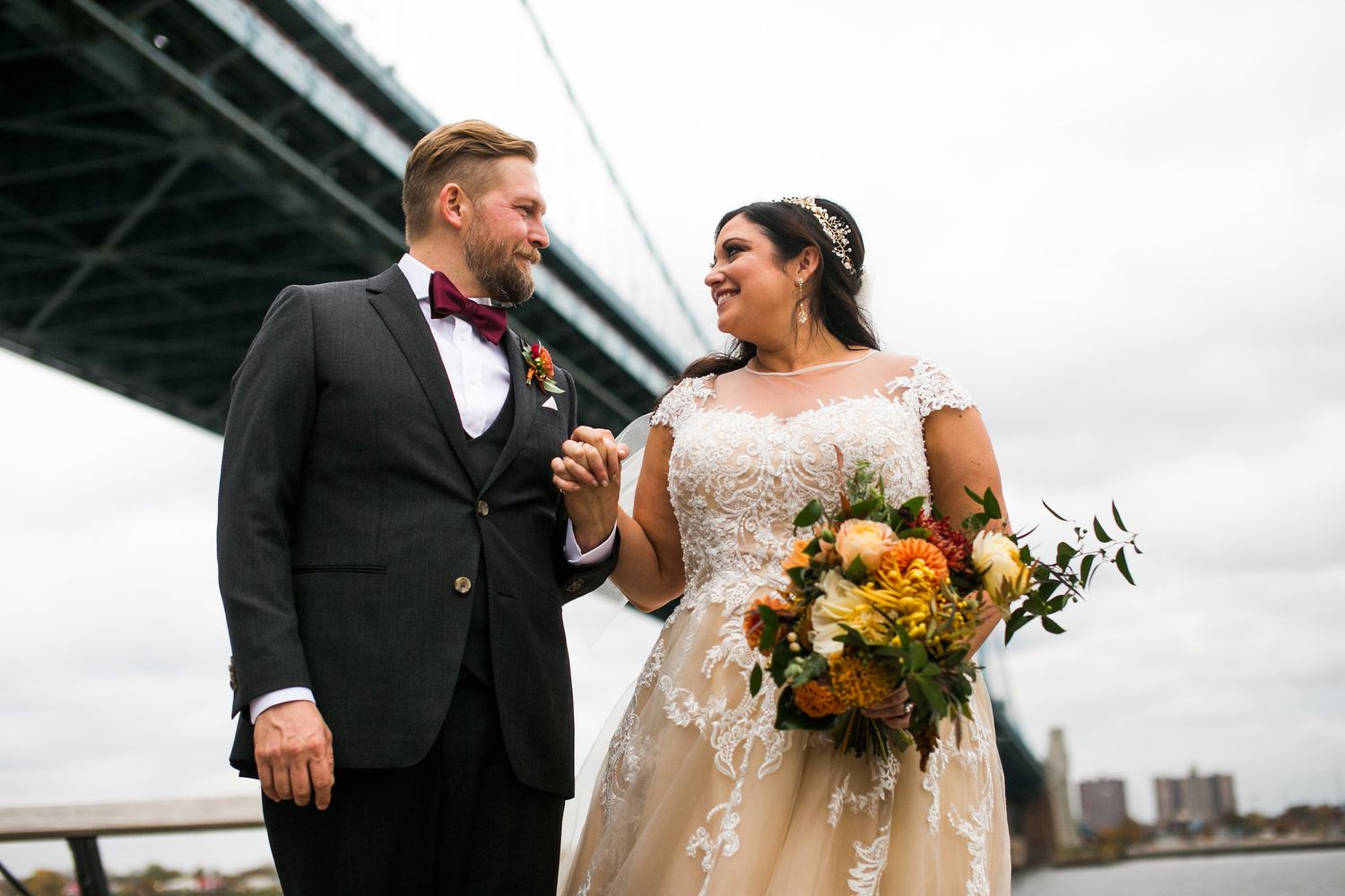 Smiling bride and groom stand together by a river bridge, bride holding a colorful fall floral bouquet.