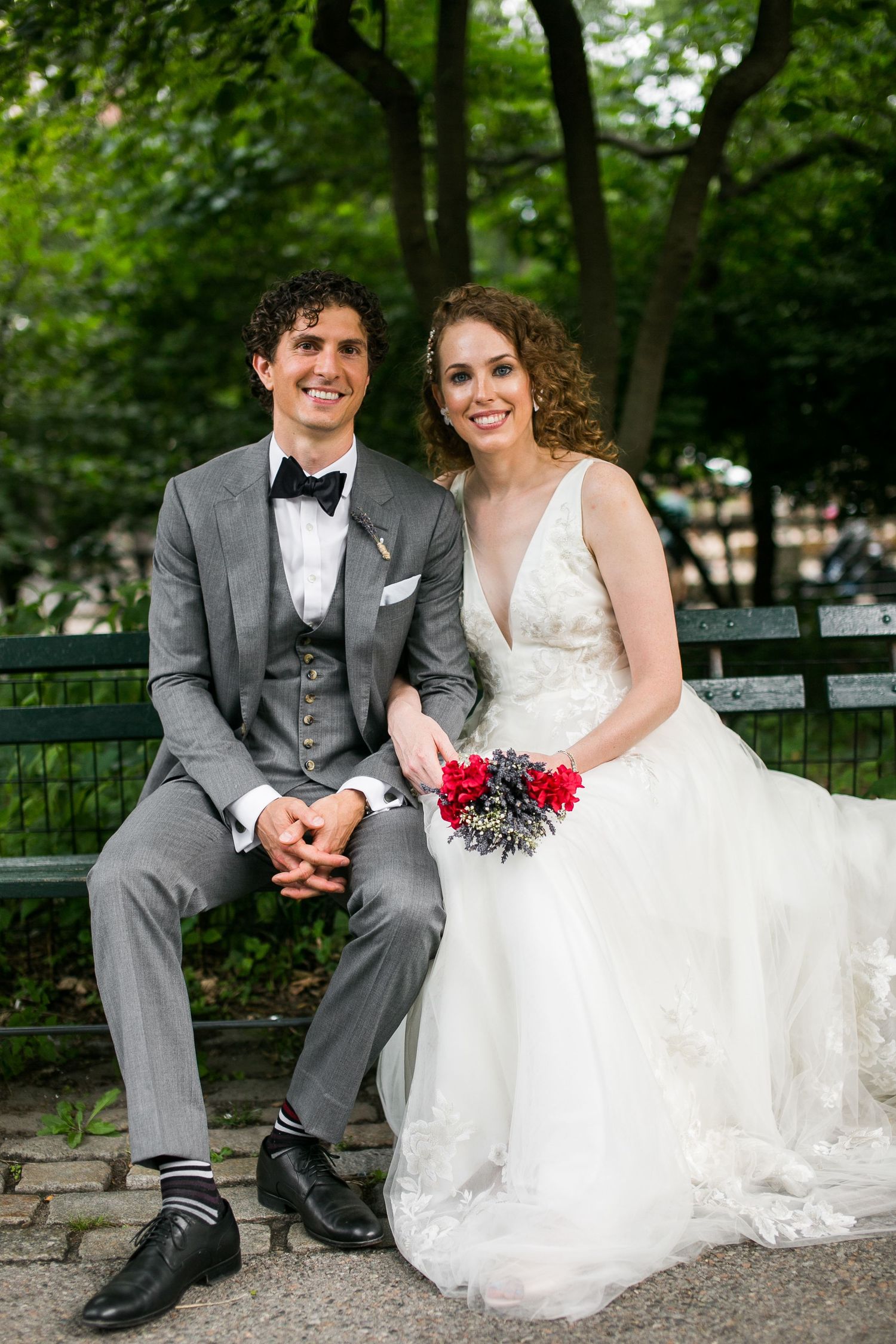 Bride and groom sitting on a green park bench, she holding red floral bouquet, both smiling at camera.