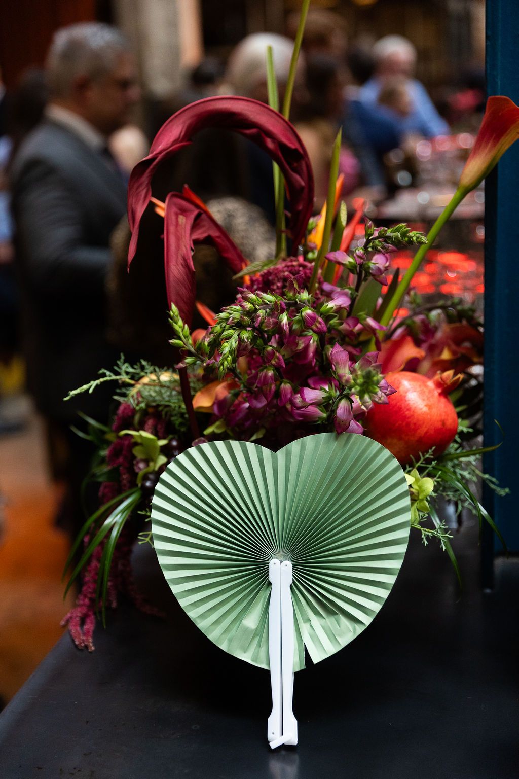 Elegant floral arrangement with burgundy blooms, pomegranate, and mint green paper fan centerpiece at an event.