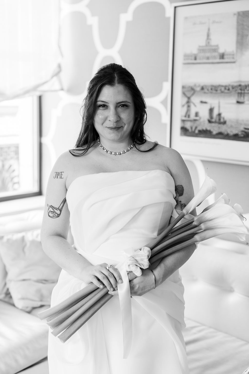 Black and white portrait of tattooed bride in strapless gown holding calla lily bouquet in hotel suite.