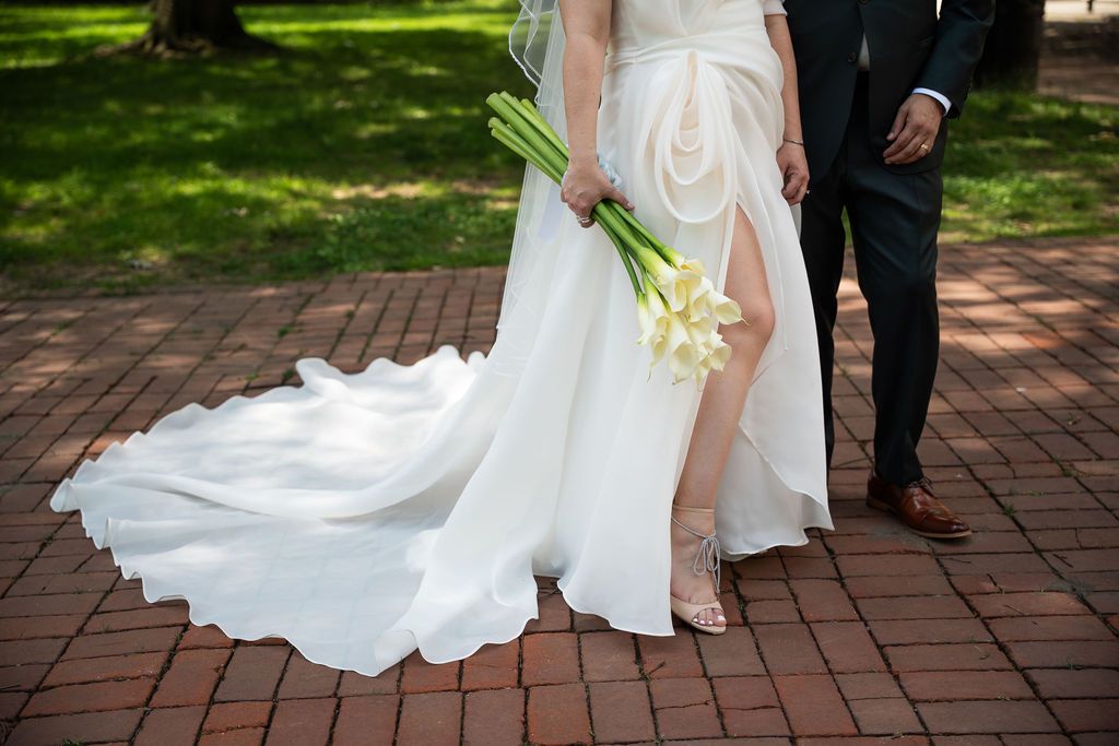 Bride in elegant white wedding gown with train holds calla lily bouquet beside groom on brick path.