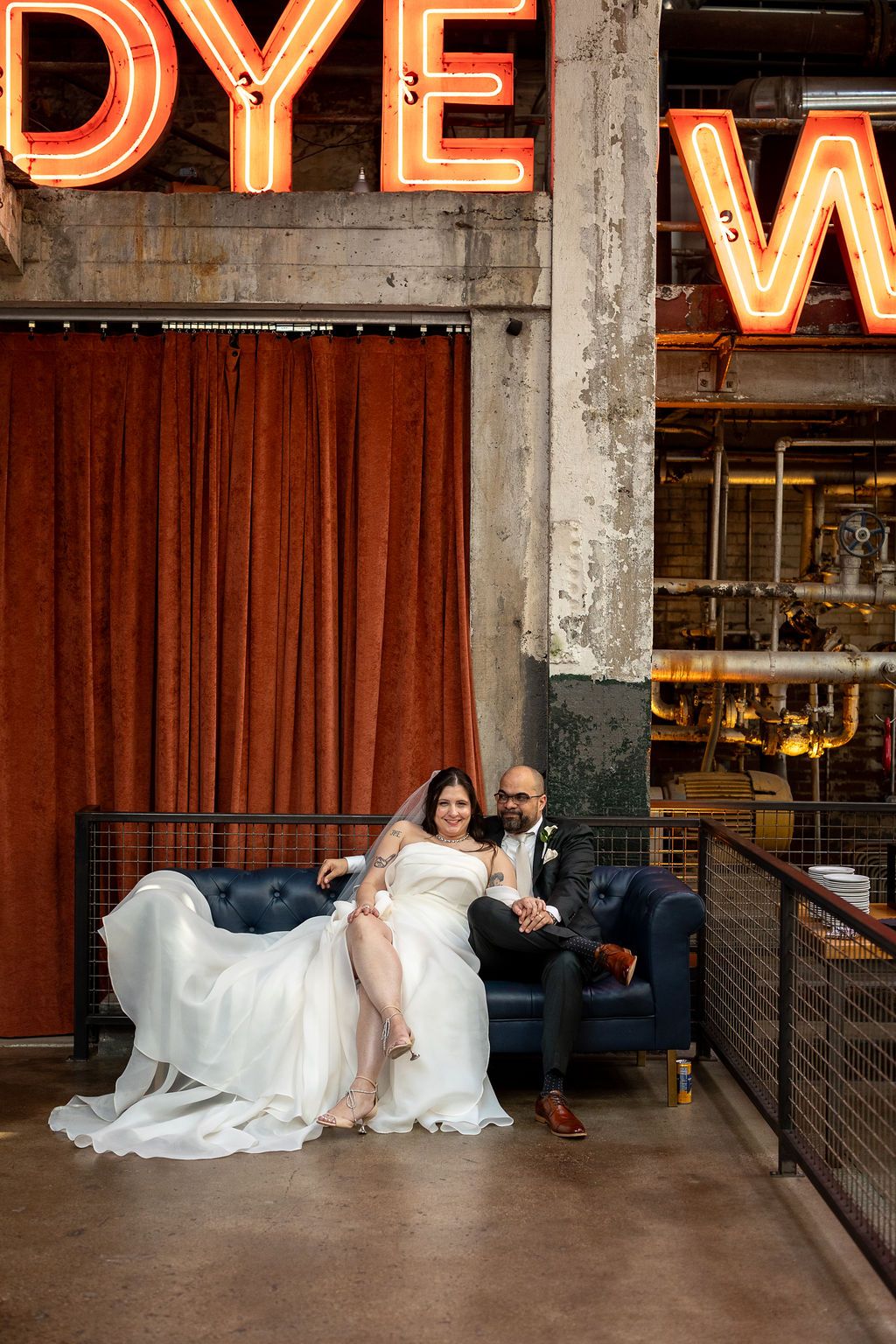 Bride and groom sitting on navy blue couch in industrial wedding venue with glowing neon signs and rust curtains.