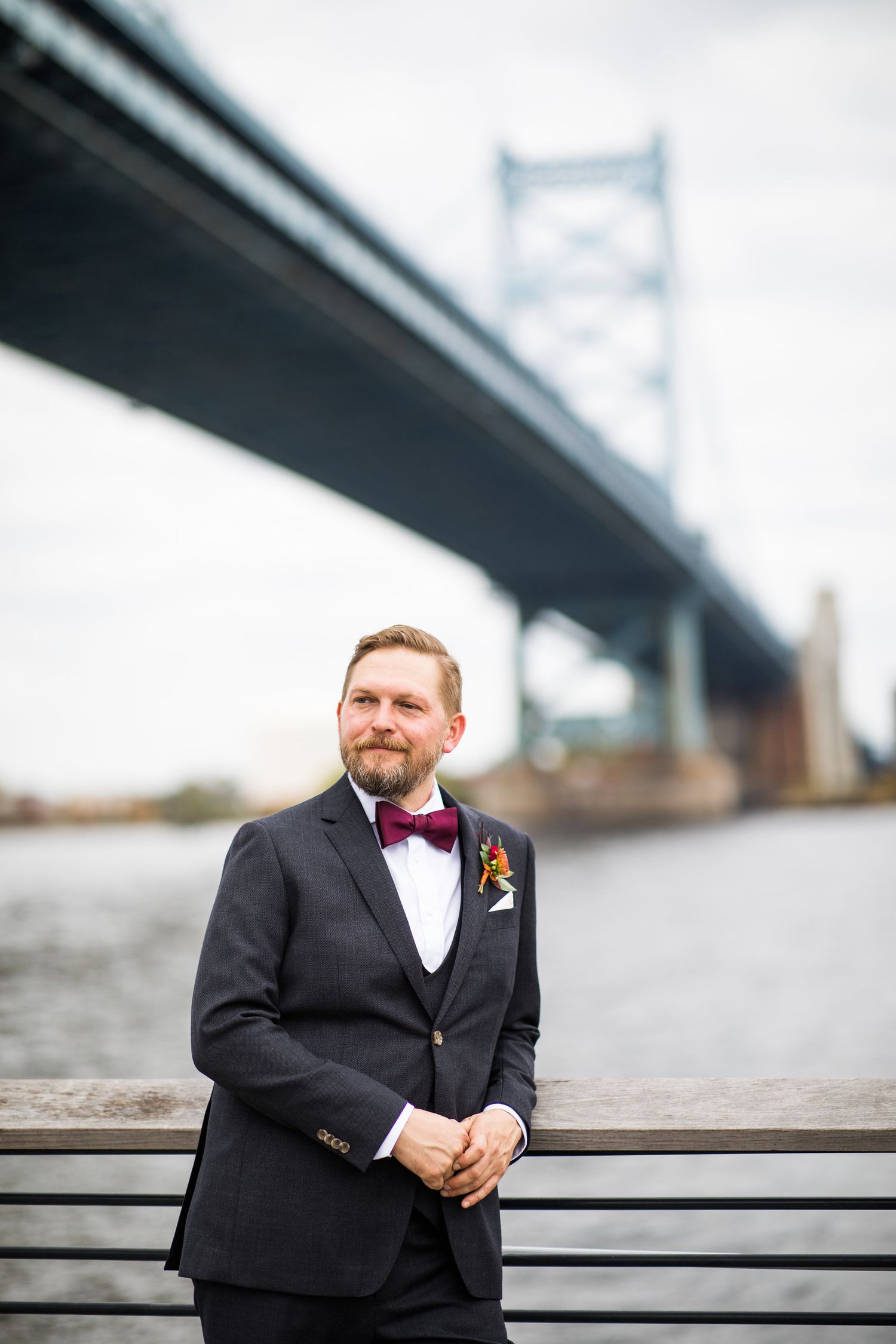 Groom in dark suit with burgundy bow tie and boutonniere posing by a river with a large suspension bridge backdrop.