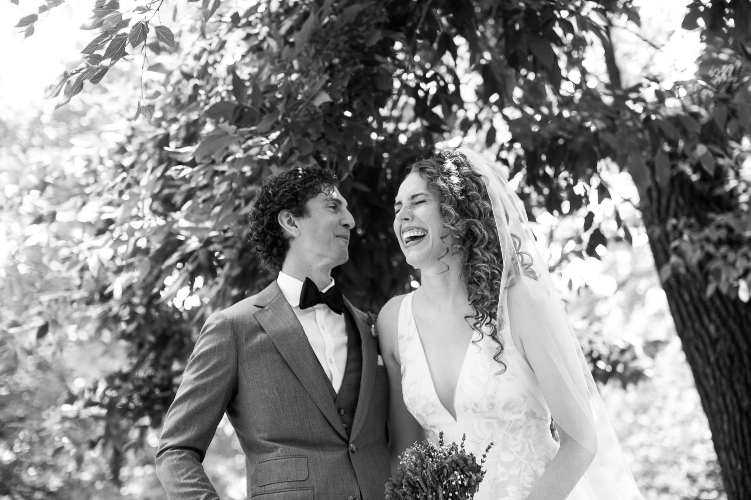 Black and white photo of laughing bride and groom under trees.