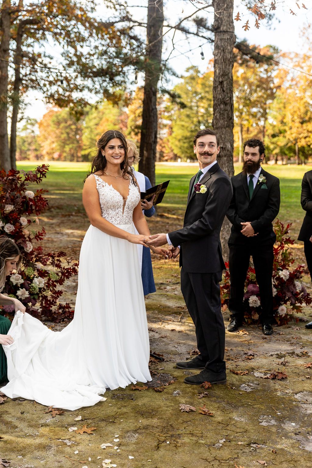 Smiling bride and groom hold hands during outdoor fall wedding ceremony with groomsmen standing behind them.