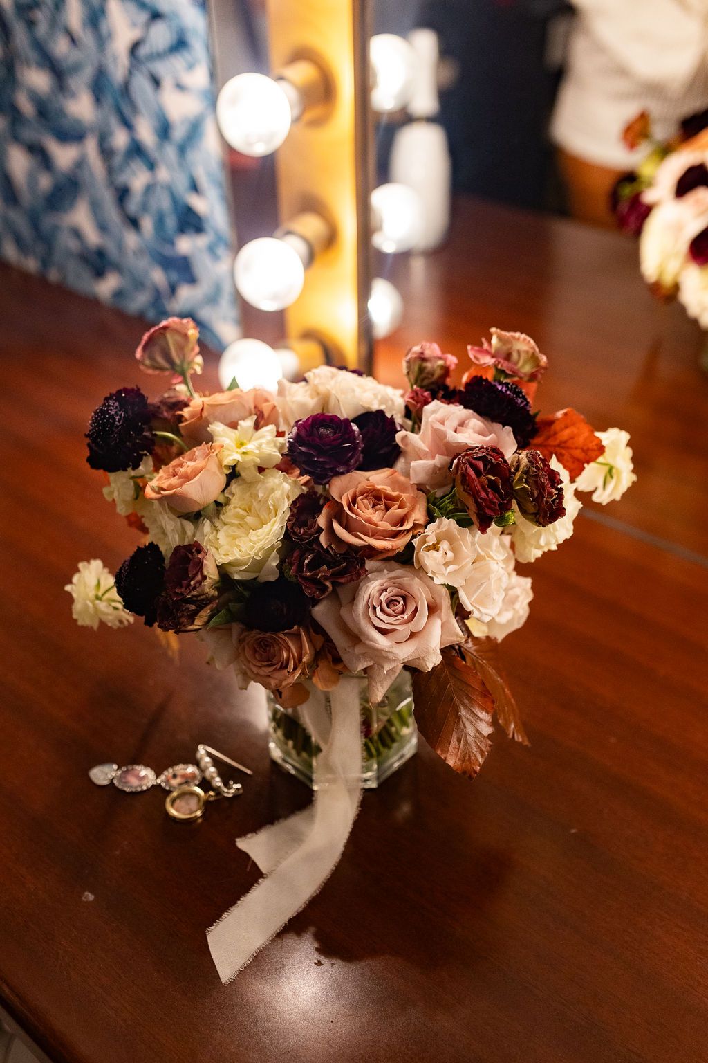 Elegant bridal bouquet with burgundy, blush, and cream roses in a glass vase on a vanity table with Hollywood mirror lights.