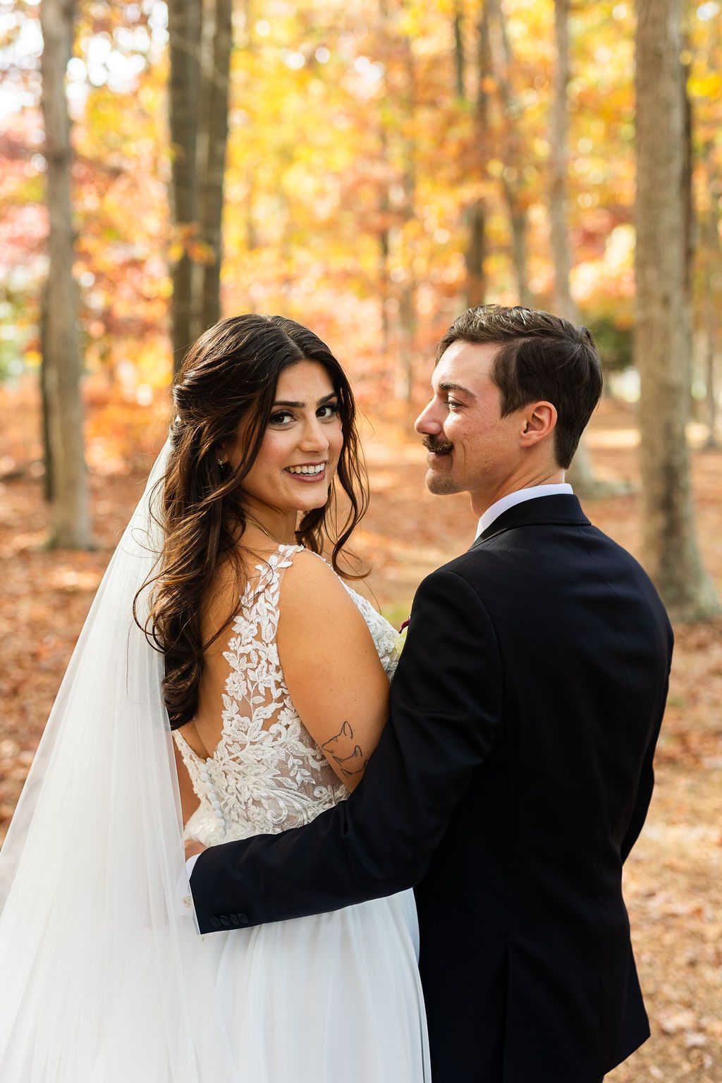 Bride in lace backless wedding gown smiling over shoulder at groom in black suit among fall foliage.