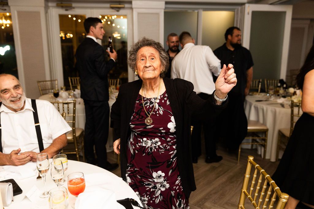 Elderly woman in a floral dress joyfully dances at a wedding reception surrounded by seated guests.