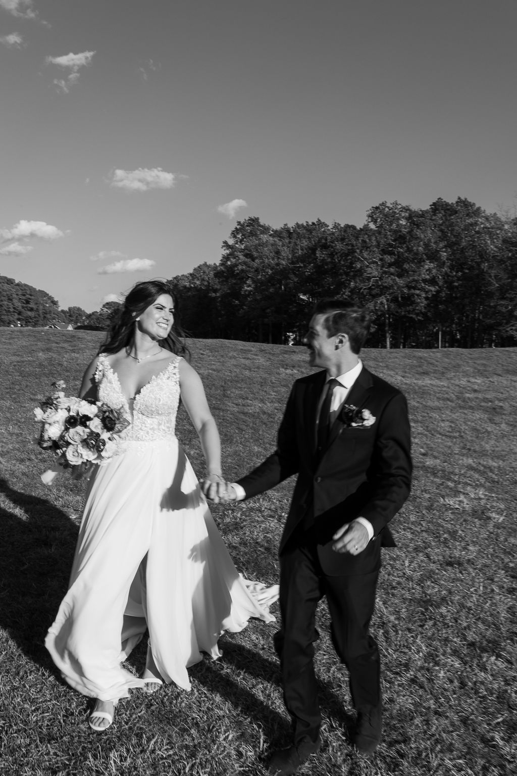 Black and white photo of bride and groom holding hands walking across sunlit grassy field with trees.