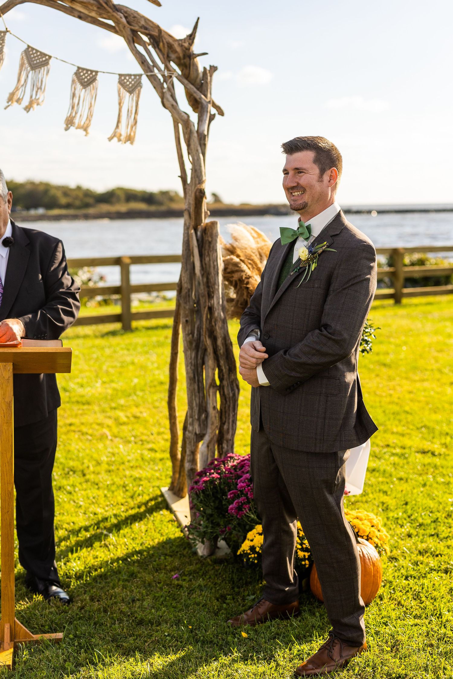 Groom in a brown plaid suit with green bow tie laughs at a driftwood altar decorated with pumpkins and mums.