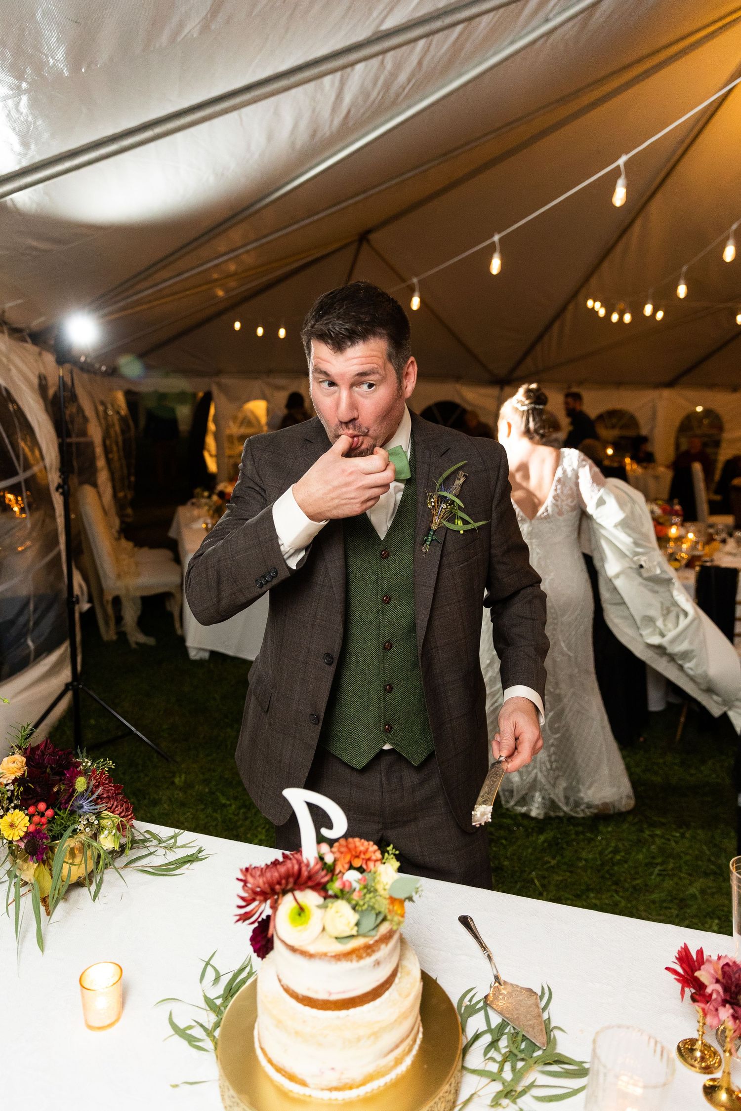 Groom tastes wedding cake while bride dances behind him at colorful tented reception celebration.