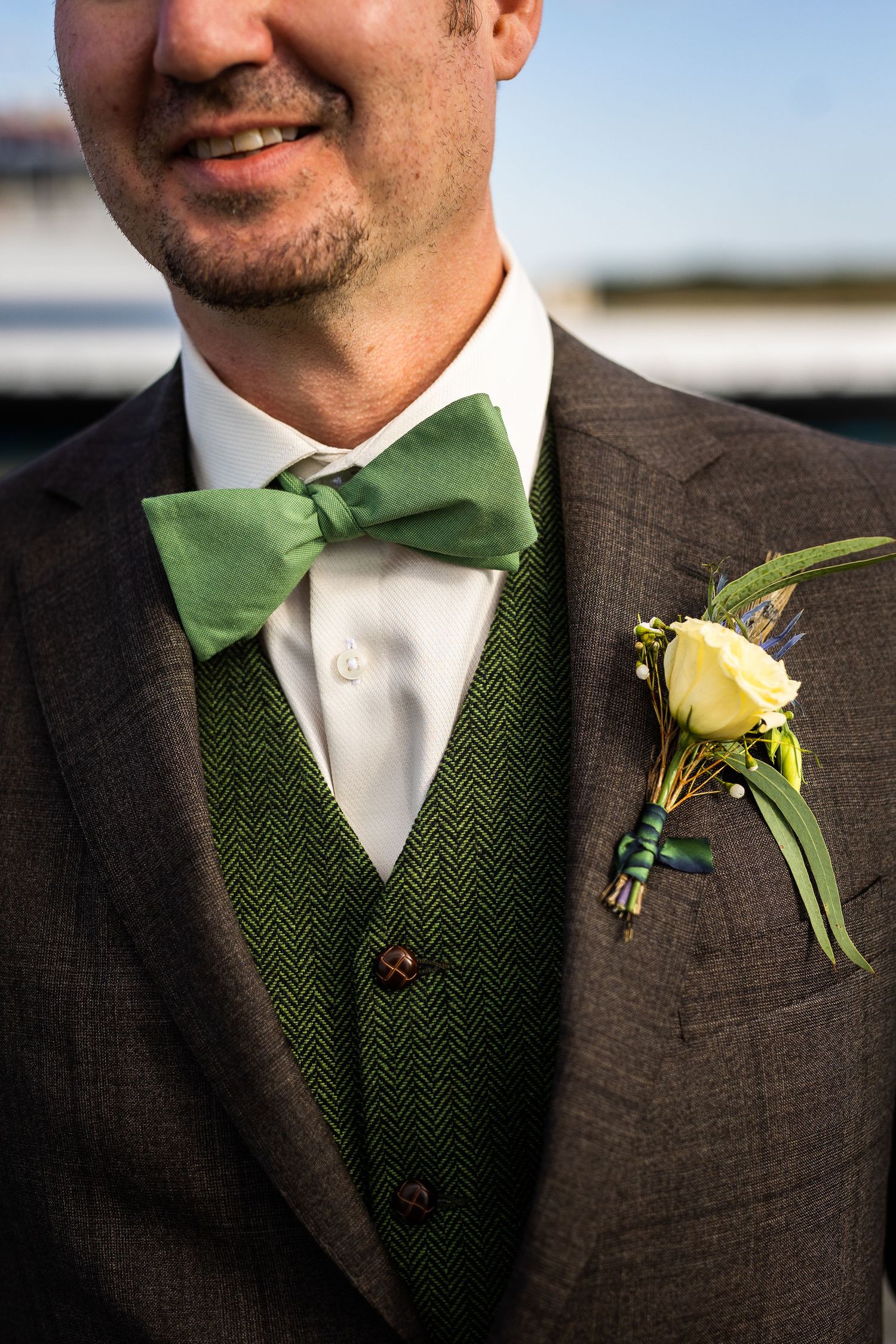 Groom in brown herringbone suit with green bow tie and yellow rose boutonniere outdoors.