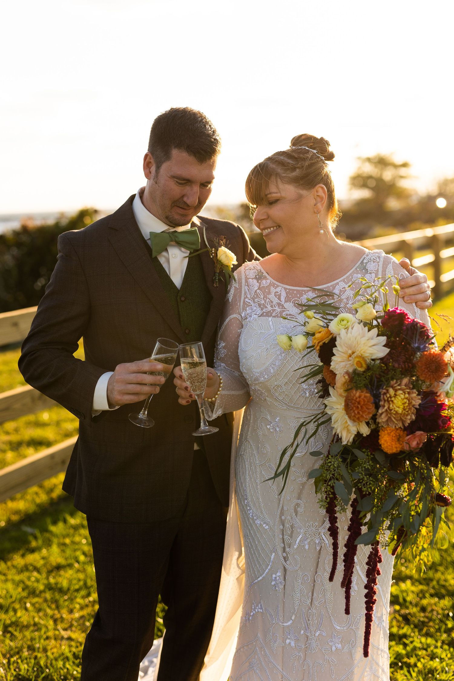 Bride and groom toasting with champagne at sunset outdoor wedding, bride holding colorful bouquet.
