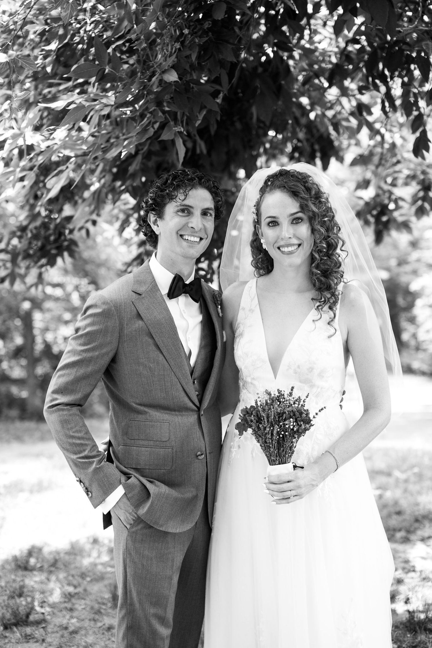 Black and white portrait of smiling bride and groom posing together under leafy tree on wedding day.