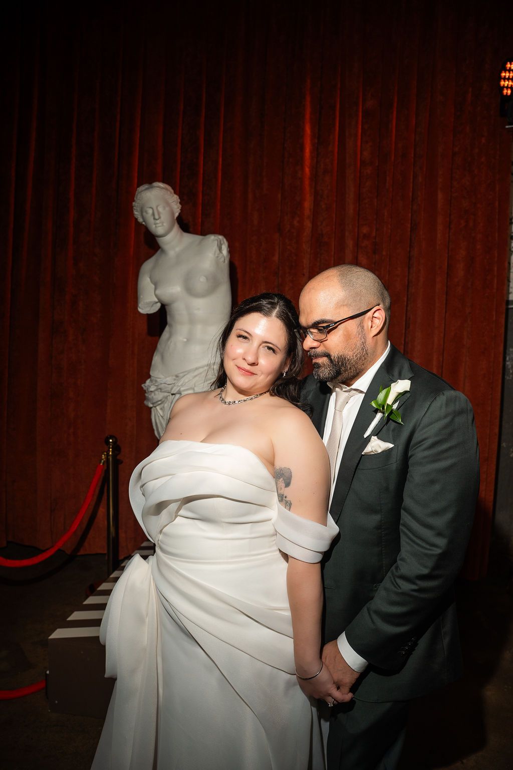 Wedding couple embracing tenderly in front of classical Venus de Milo statue and red curtain.