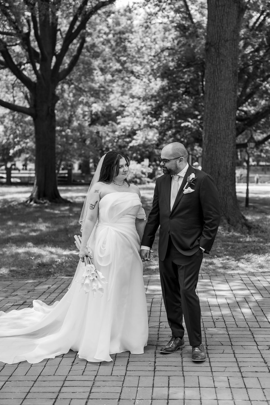 Black and white photo of bride and groom holding hands walking on brick path among tall park trees.