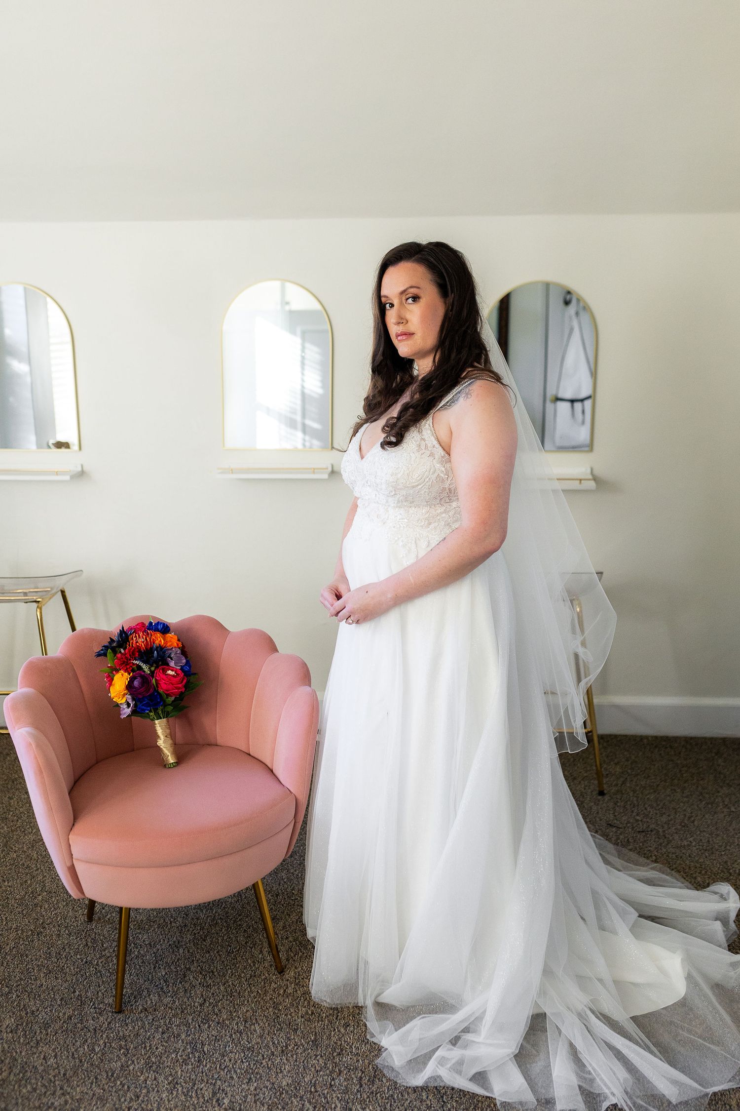 Bride in white lace wedding gown standing beside pink velvet chair with colorful bouquet.