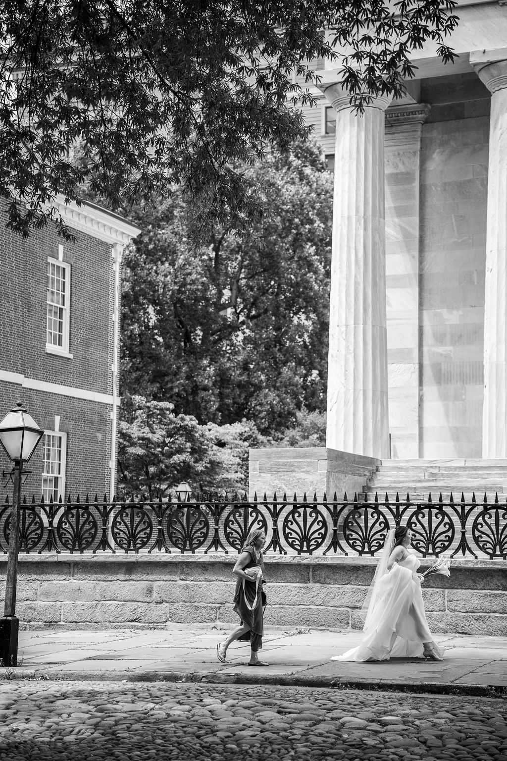 Black and white photo of a bride in a white gown walking past ornate iron fence near historic building.