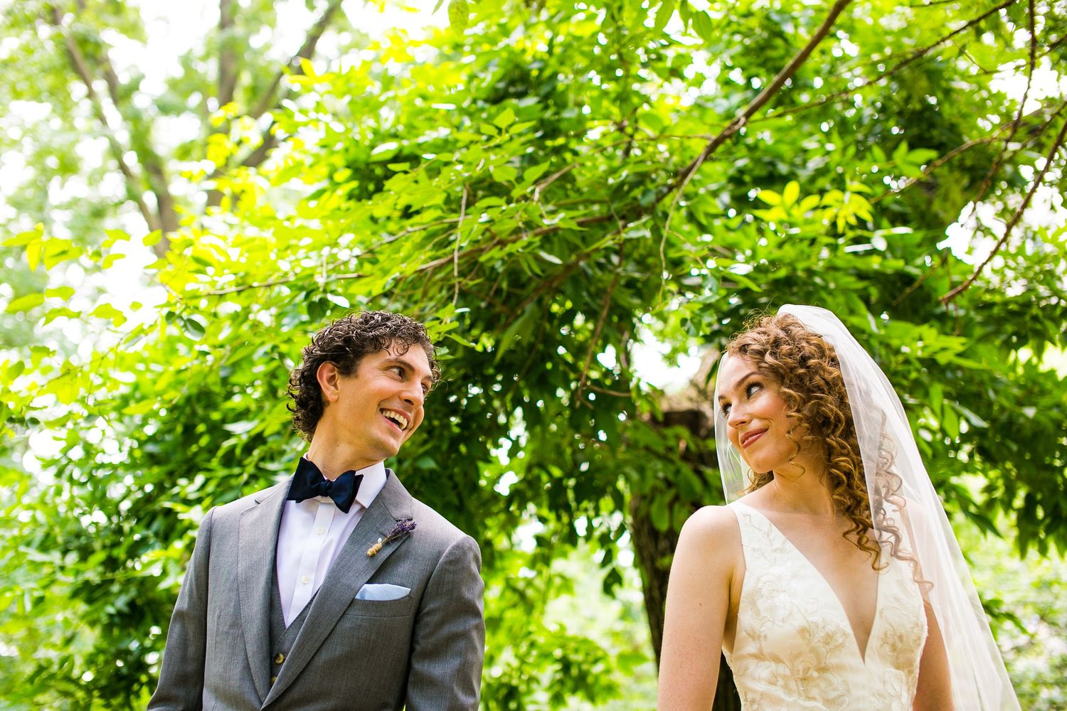 Bride and groom share a joyful glance surrounded by lush green trees on their wedding day.