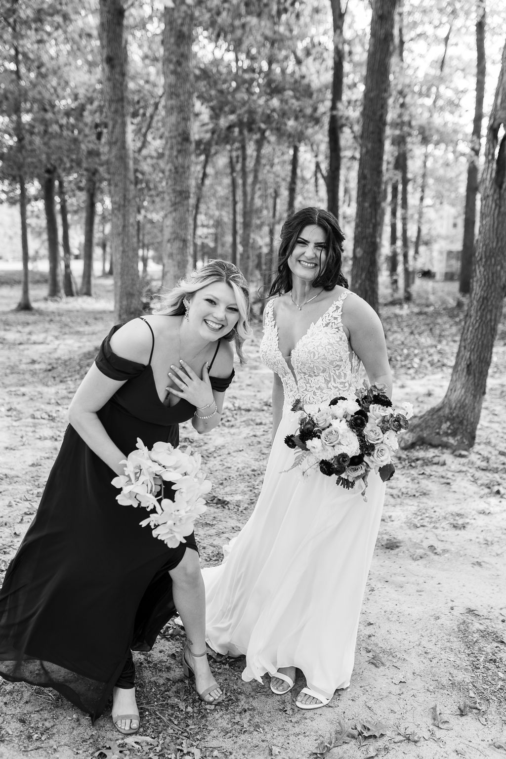 Laughing bride and bridesmaid posing playfully in a forest setting in a black and white wedding photo.