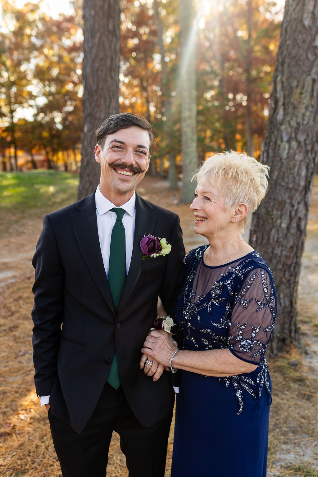Smiling groom with mustache in black suit poses with older woman in blue dress at fall wedding.