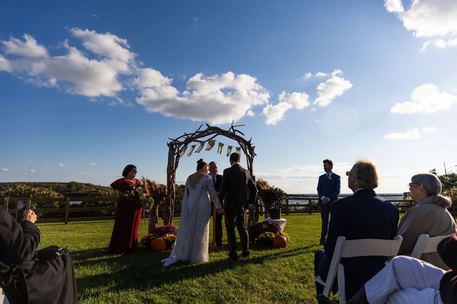 Outdoor wedding ceremony under rustic wooden arch with water view and blue sky backdrop.
