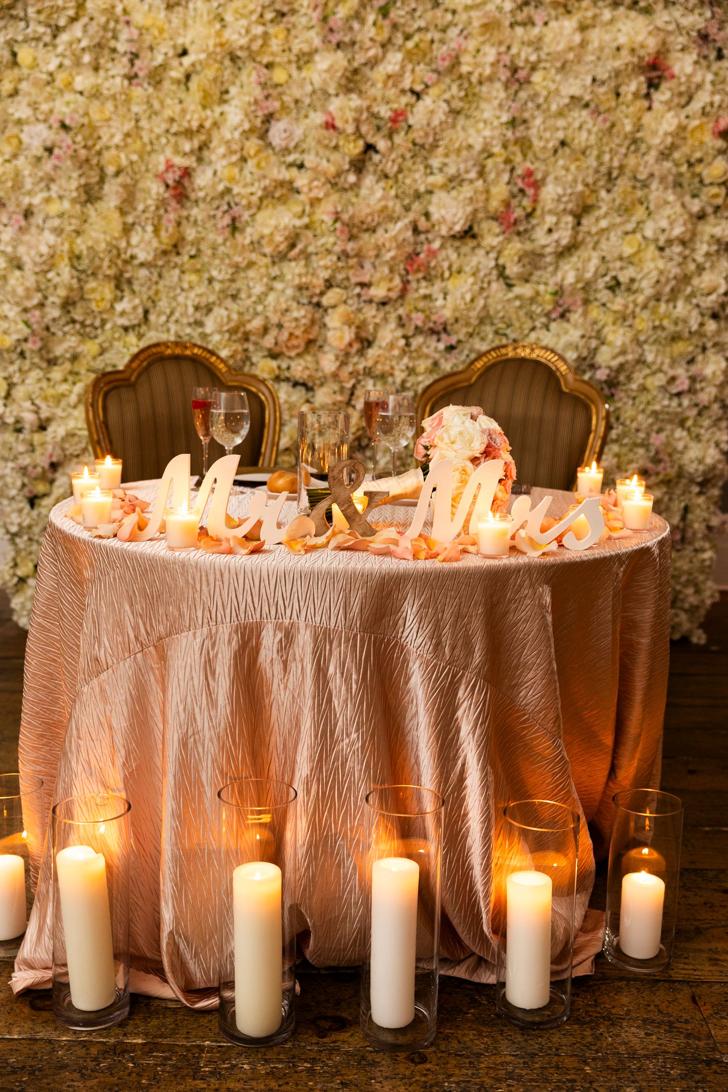 Elegant wedding sweetheart table with pink linens, candles, and a floral wall backdrop.