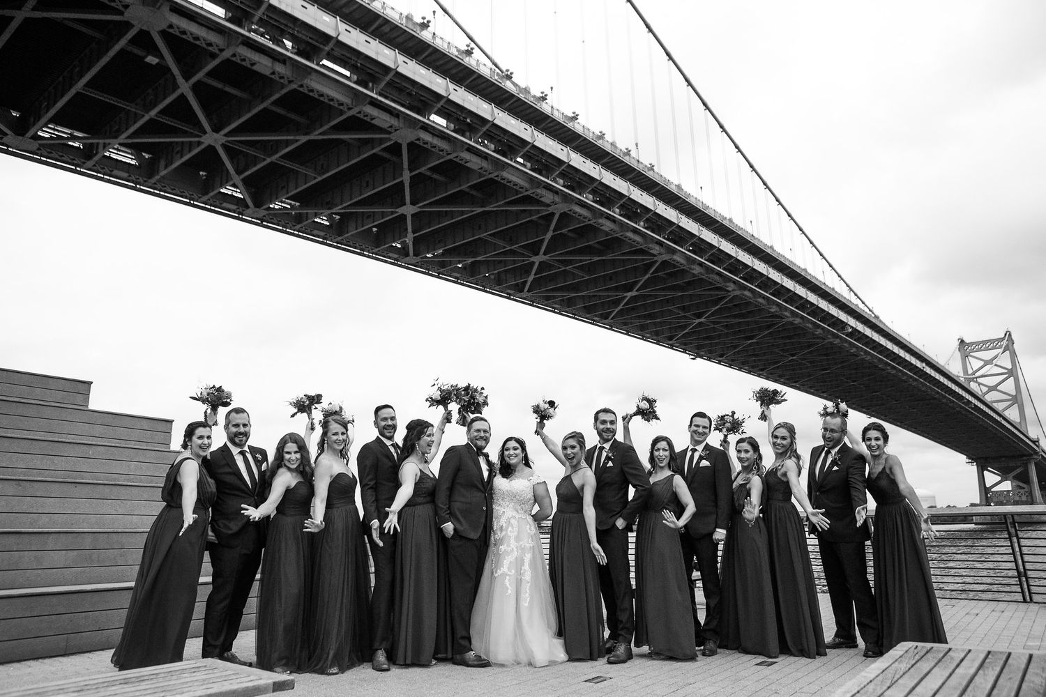 Black and white photo of large wedding party gathered under the Benjamin Franklin Bridge on a riverside deck.