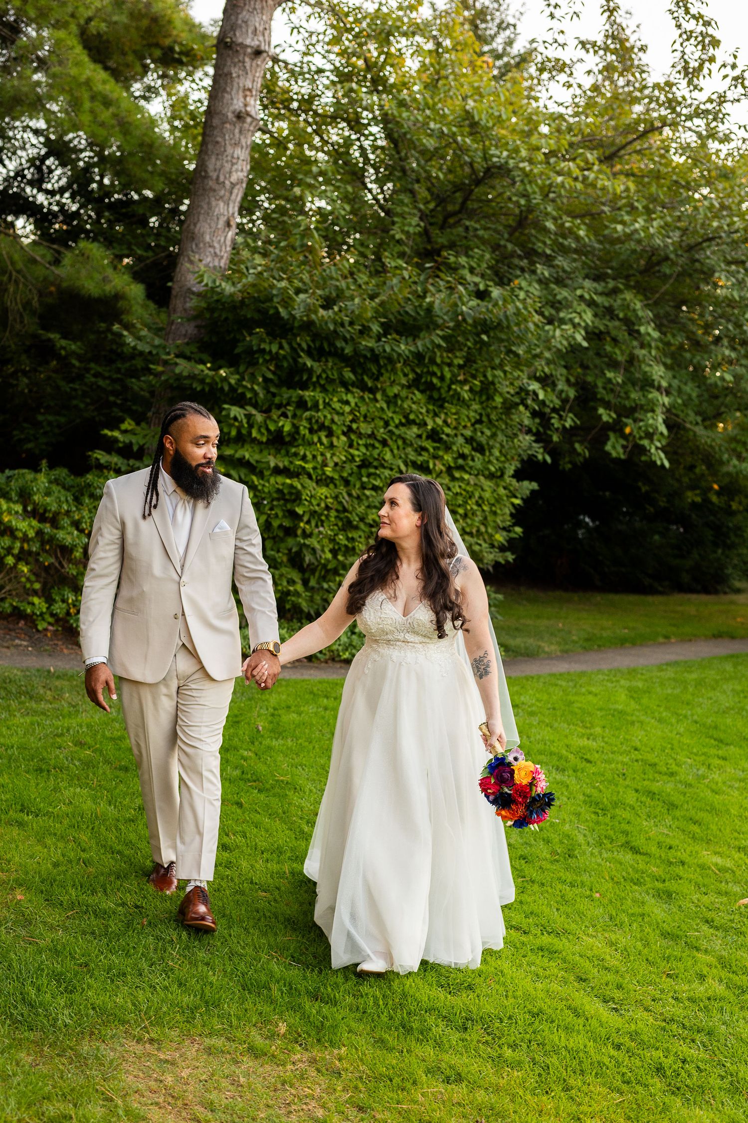Bride and groom walk hand in hand across green lawn, bride holding vibrant colorful wedding bouquet.