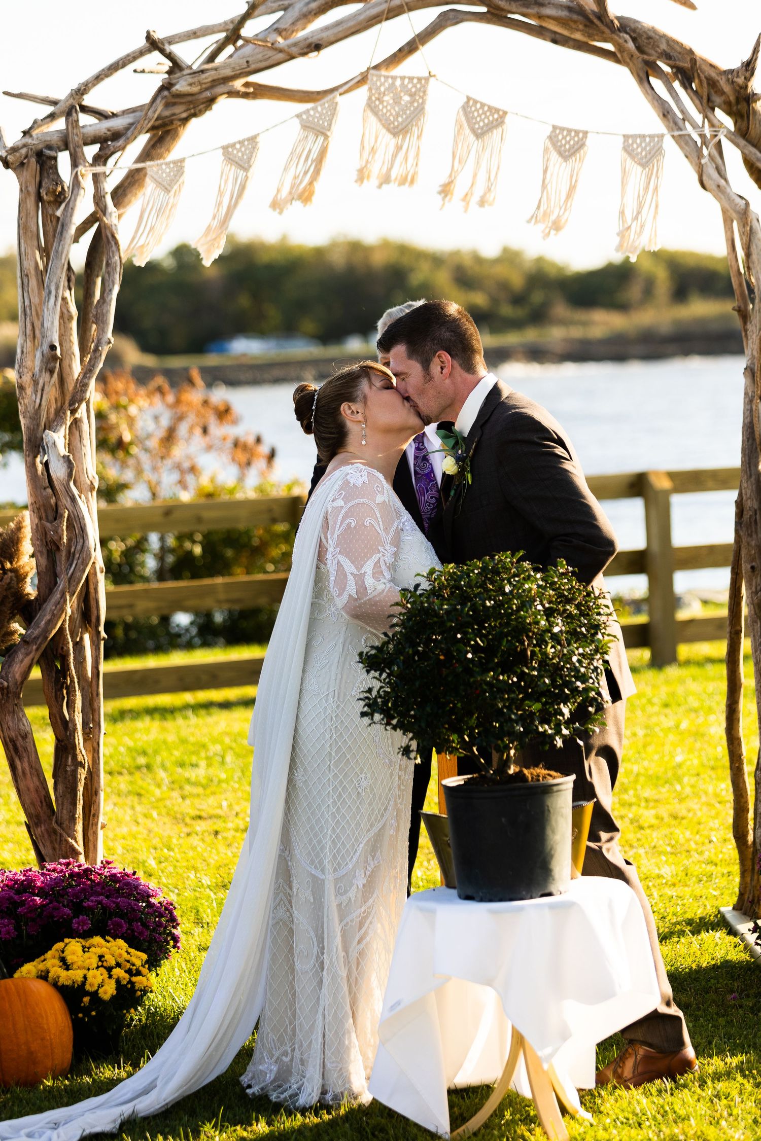 Bride and groom sharing first kiss under rustic driftwood arch with macrame bunting by waterfront.