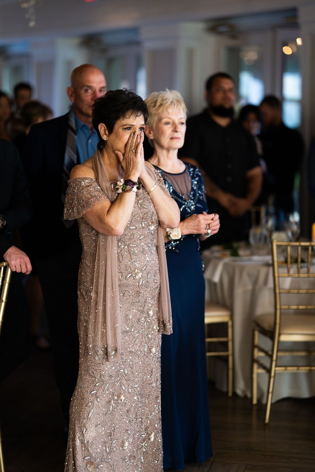 Emotional mother wipes tears watching wedding first dance, standing beside woman in navy blue formal gown.